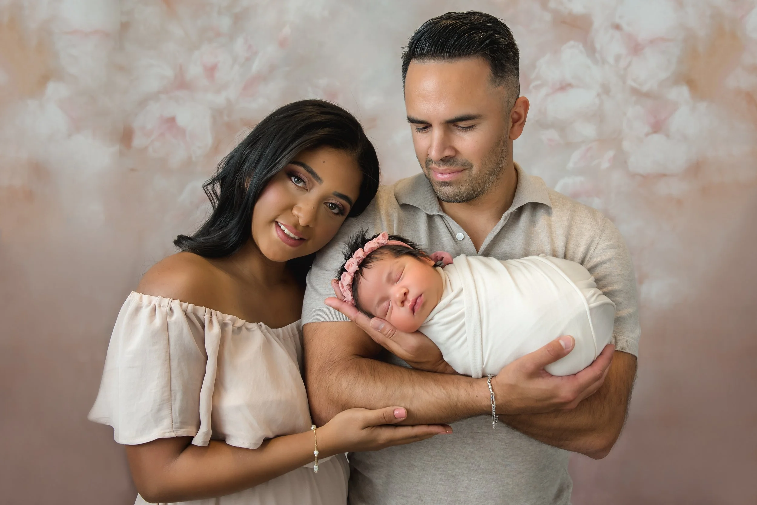 A happy family of three with a pregnant woman, her partner, and their newborn baby girl, who is swaddled in white and wearing a pink floral headband.