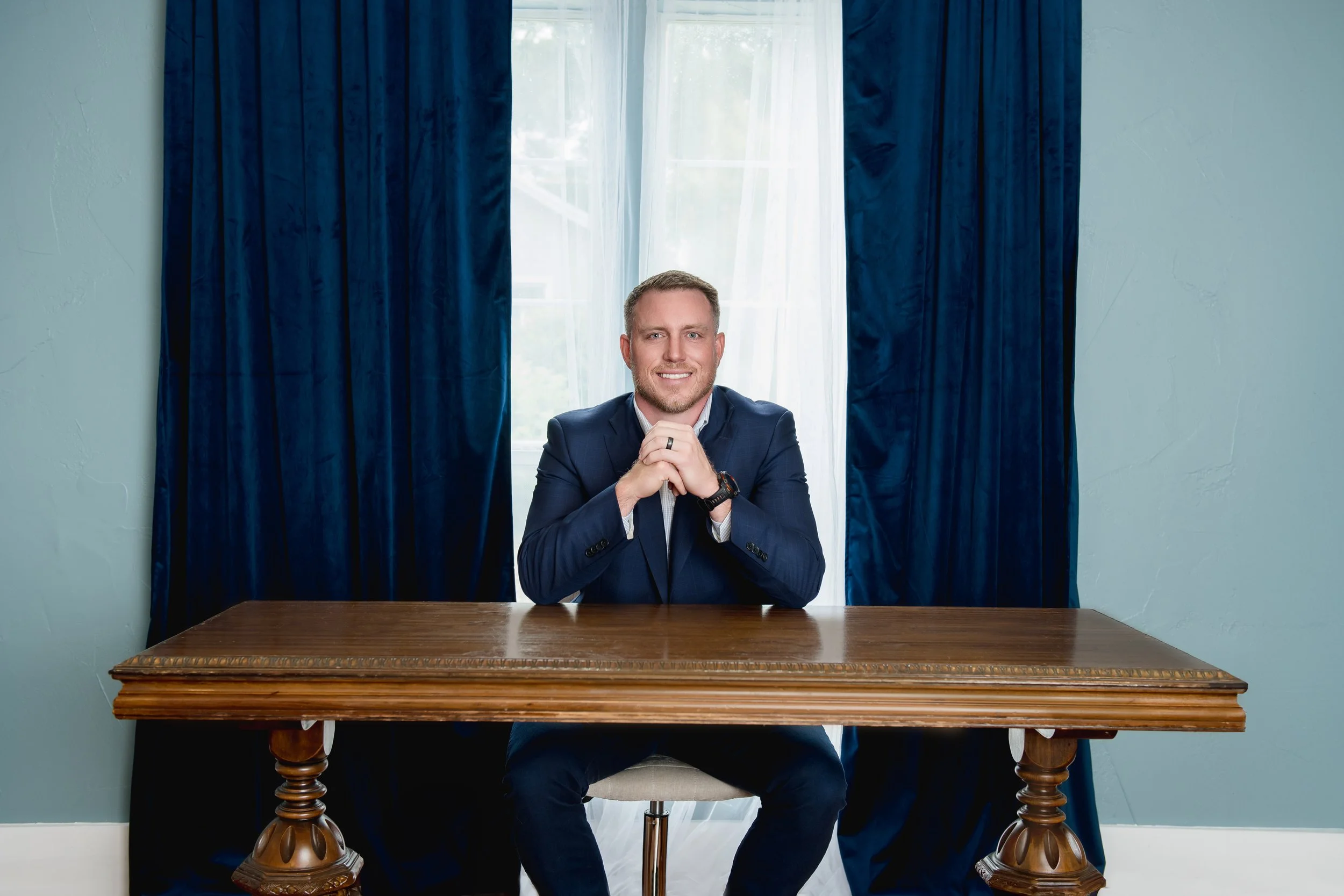 A man in a dark suit sitting at a wooden table with blue curtains and a window behind him, smiling with hands clasped.