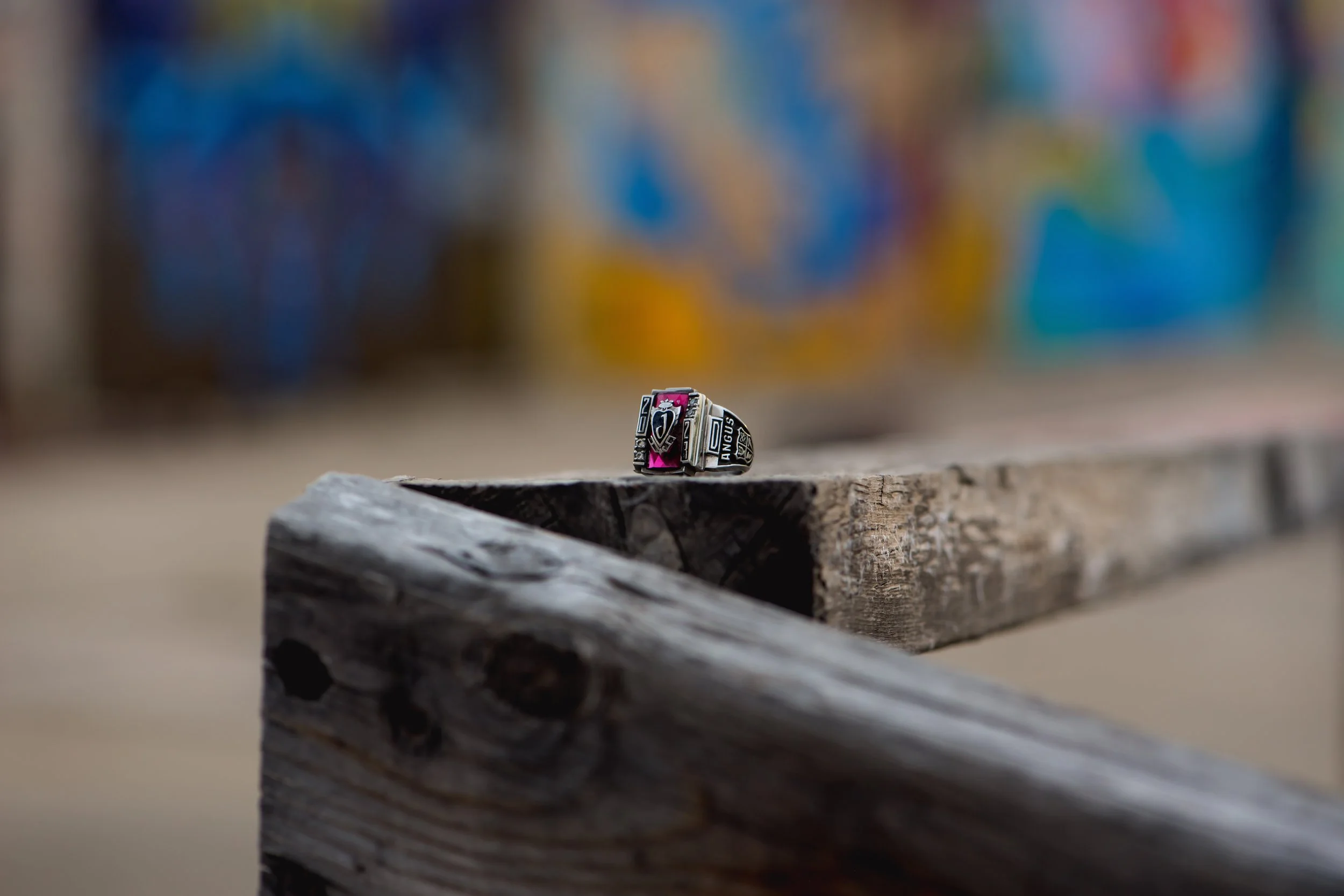A close-up of a silver ring with black and pink accents resting on a weathered wooden surface, with colorful graffiti in the blurry background.