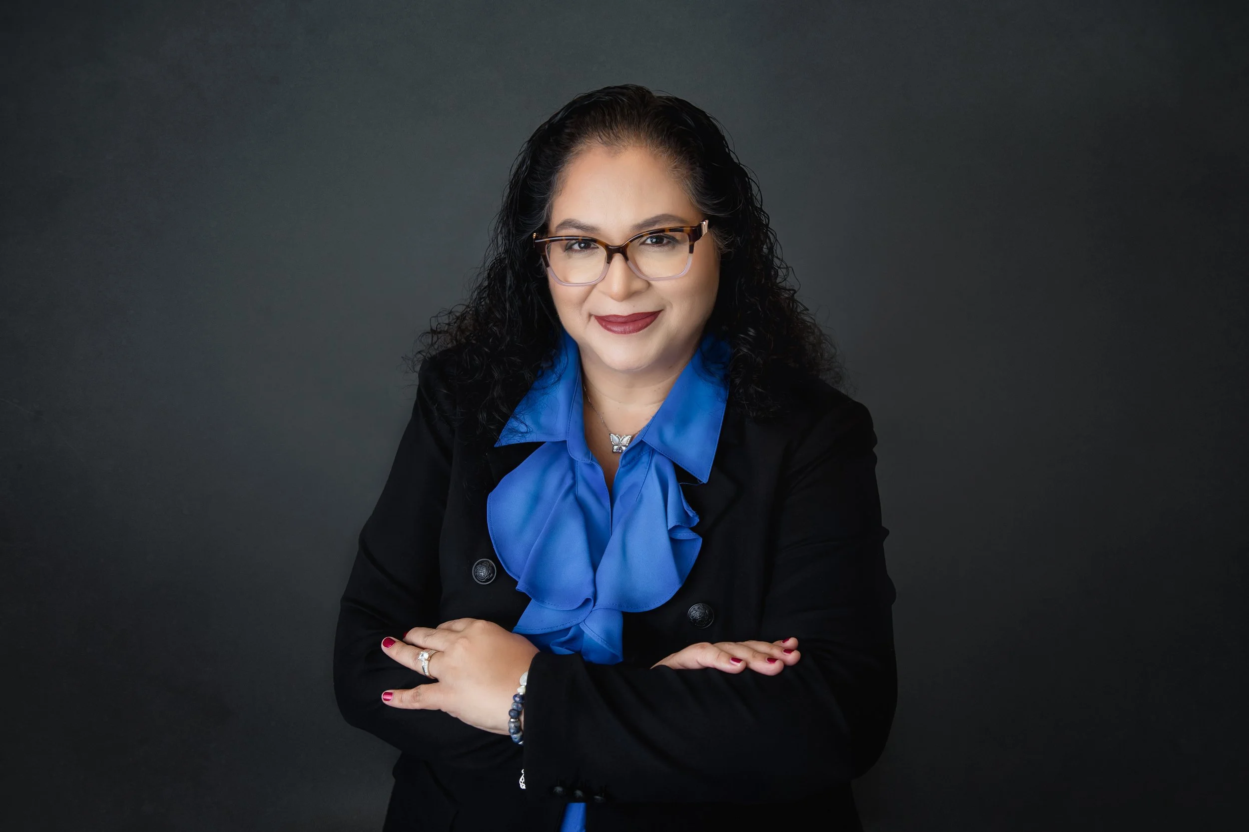 A woman with dark curly hair and glasses, wearing a blue blouse and black blazer, smiling with arms crossed against a dark background in our Spring Branch studio.