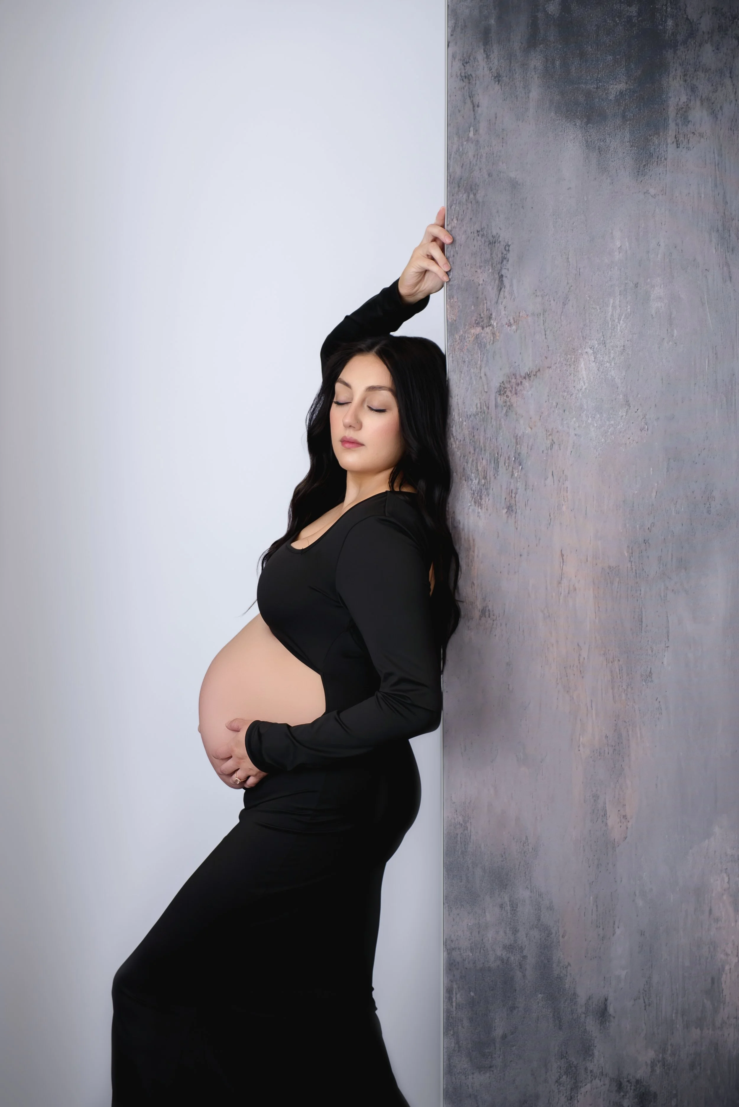 Luxury maternity portrait of a mother in a black gown leaning against a textured wall with eyes closed, hands gently resting on her baby bump.
