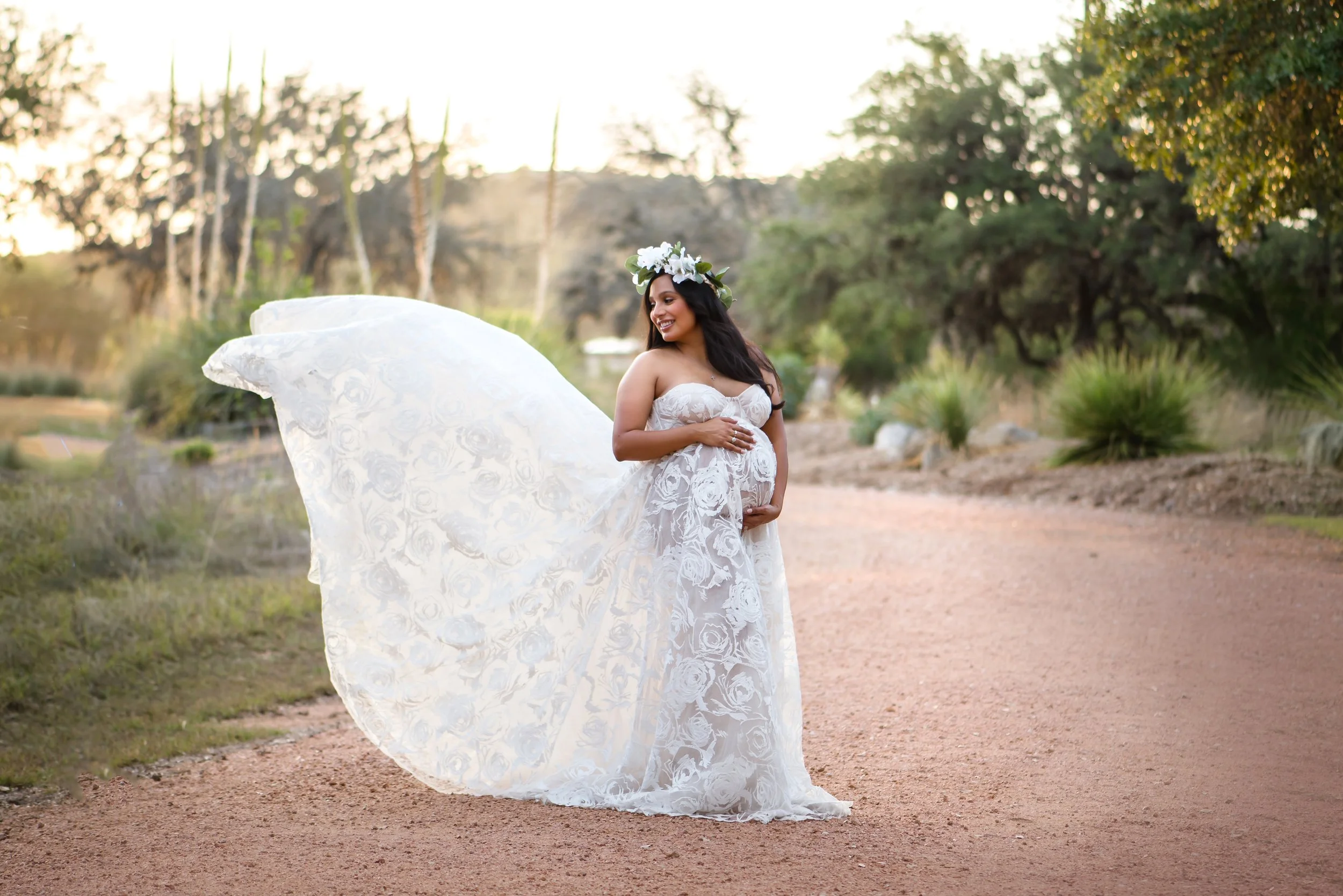 Heirloom maternity portrait of a mother in a white lace gown with floral crown, standing on a sunset-lit path in the Hill Country near Spring Branch.