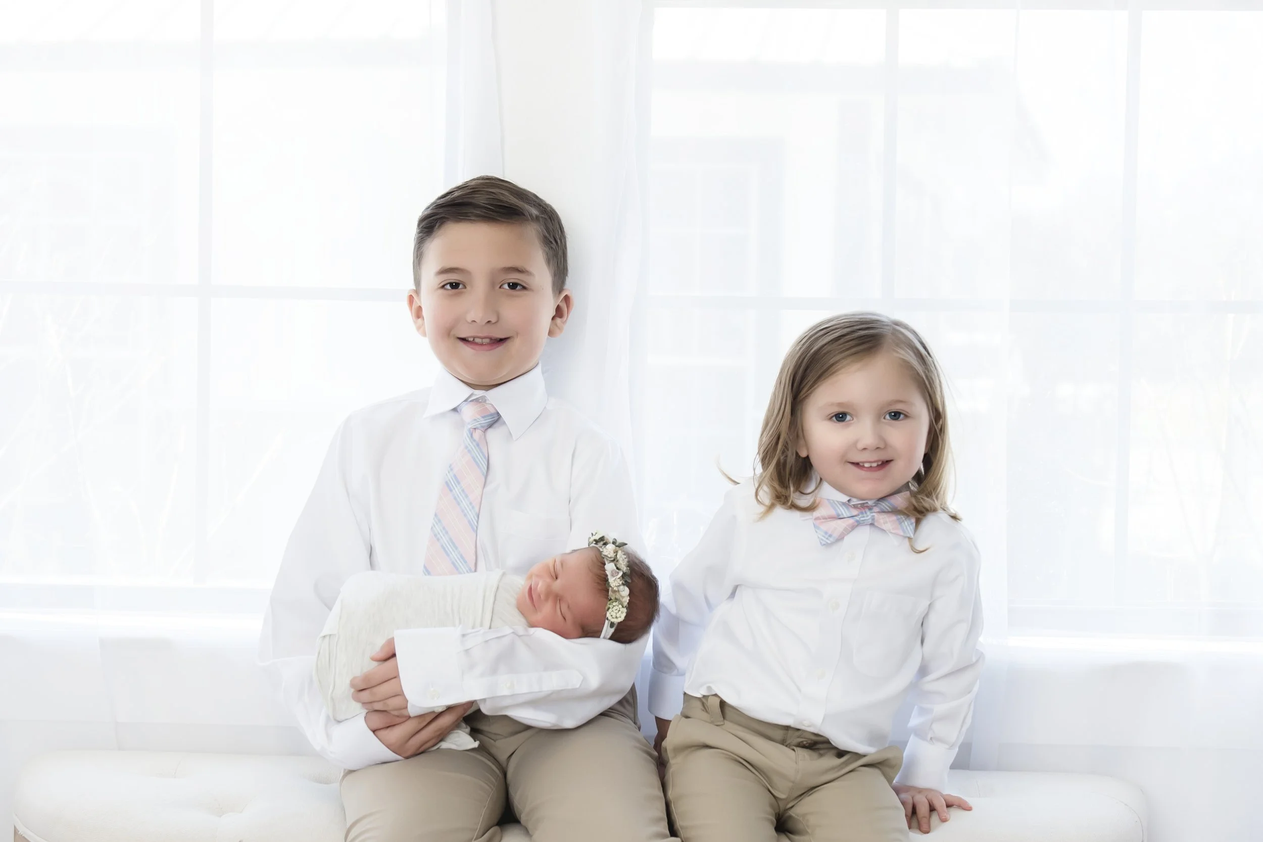 Three children, a boy, a girl, and a baby, sitting together on a white couch in a bright room with large windows. The boy is holding the baby, and all are dressed in white shirts and khaki pants, with the girl wearing a bow tie.