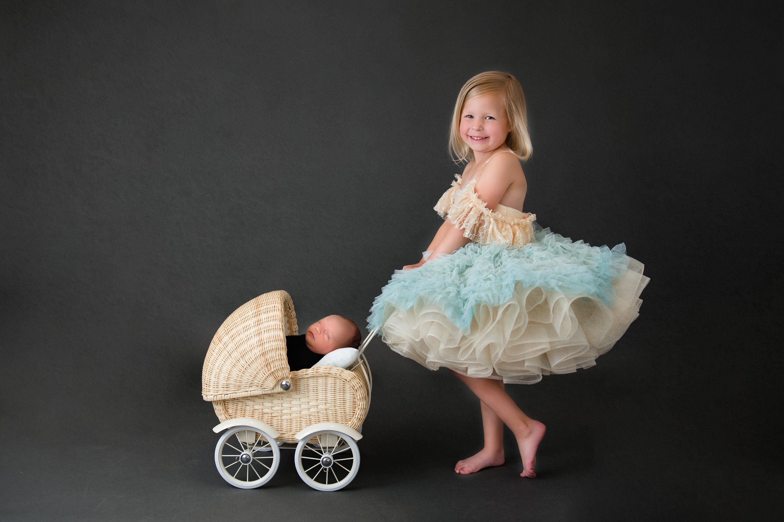 A young girl in a pastel-colored dress with a ruffled skirt standing next to a toy baby stroller with a sleeping baby inside, set against a dark background.