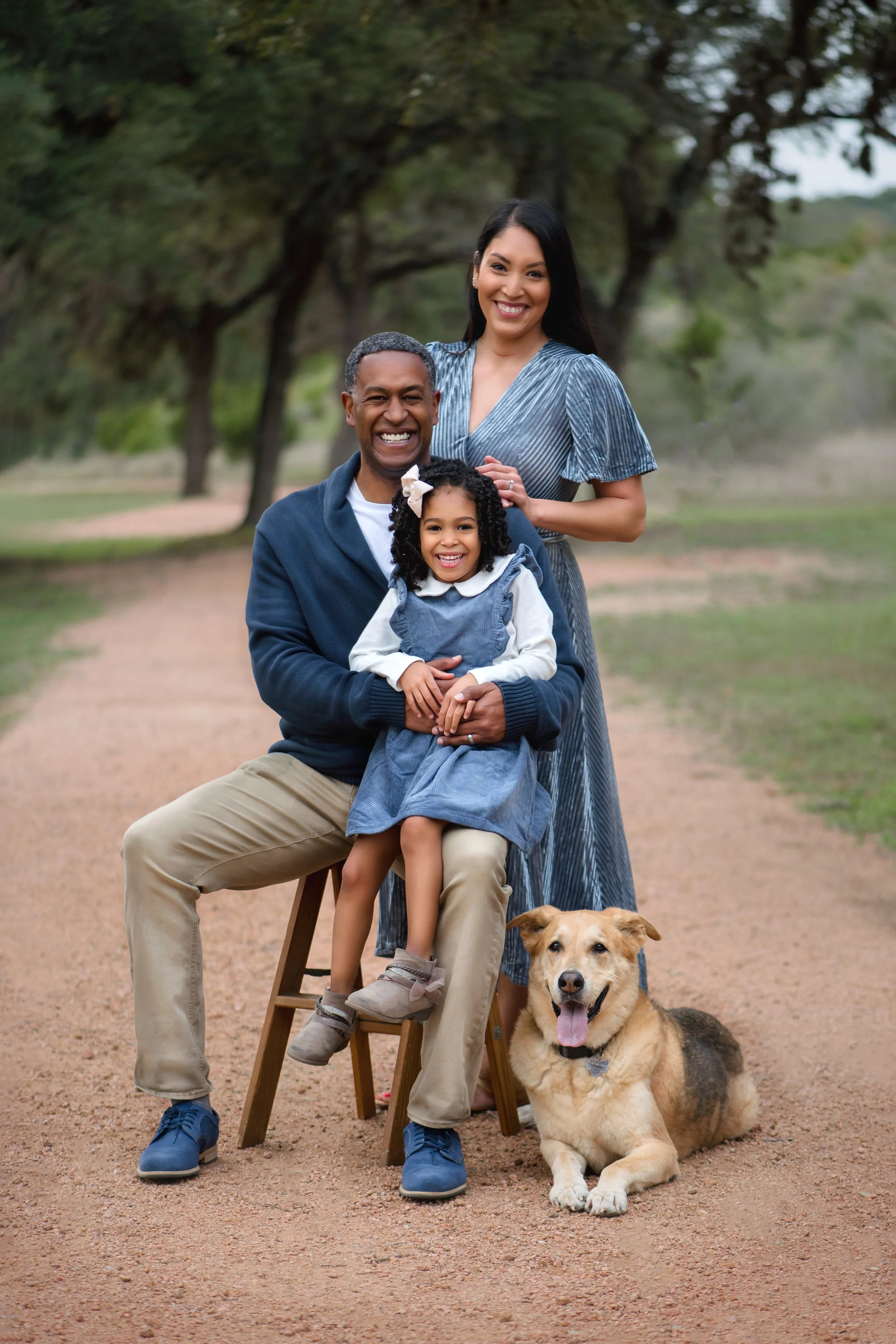 Smiling family of three and a dog outdoors on a dirt path, with trees in the background. The father is sitting on a stool, holding their young daughter, while the mother stands behind them. The dog is lying on the ground in front.
