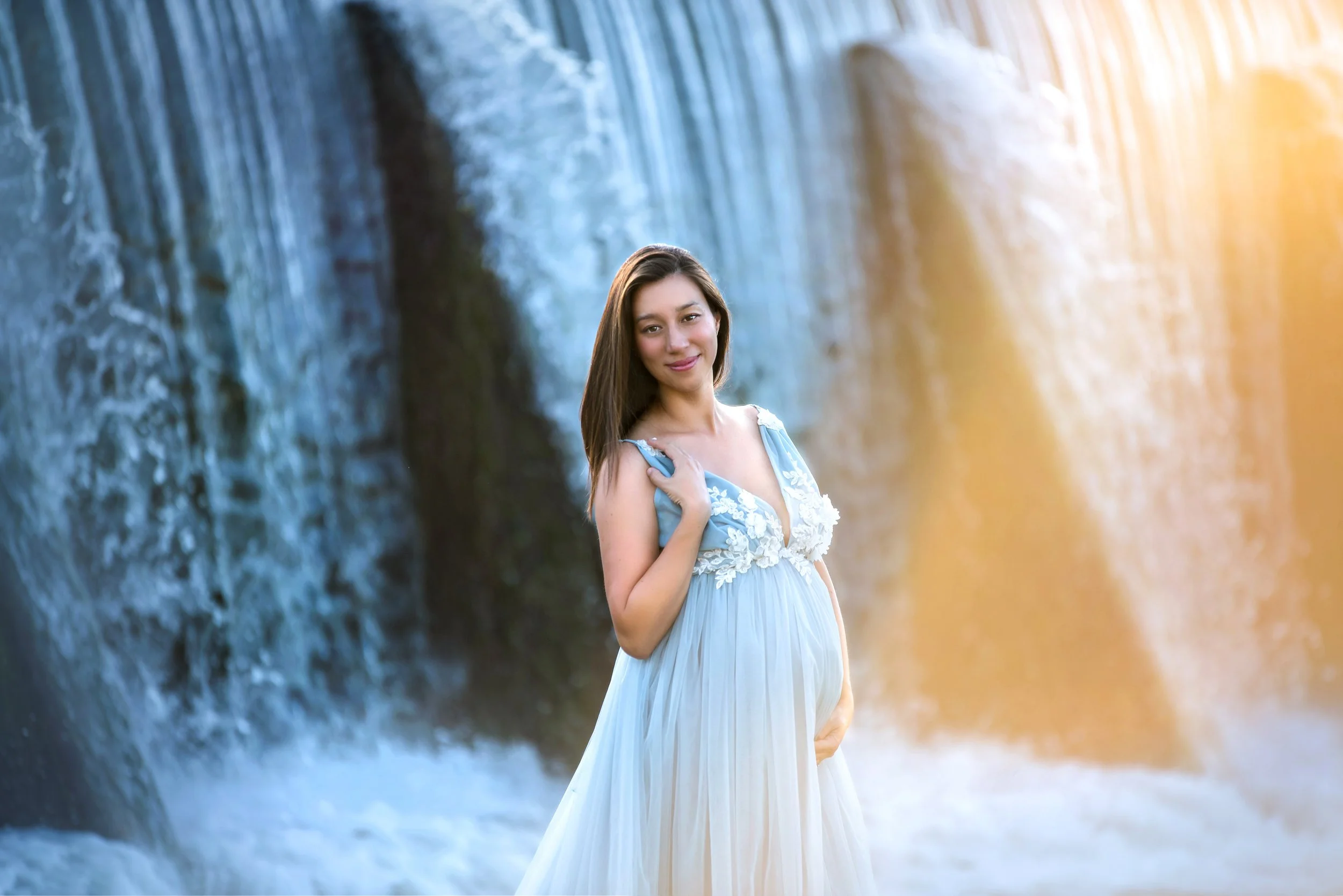 Luxury outdoor maternity portrait of a pregnant woman in a light blue dress standing before a waterfall at sunset in the Texas Hill Country.