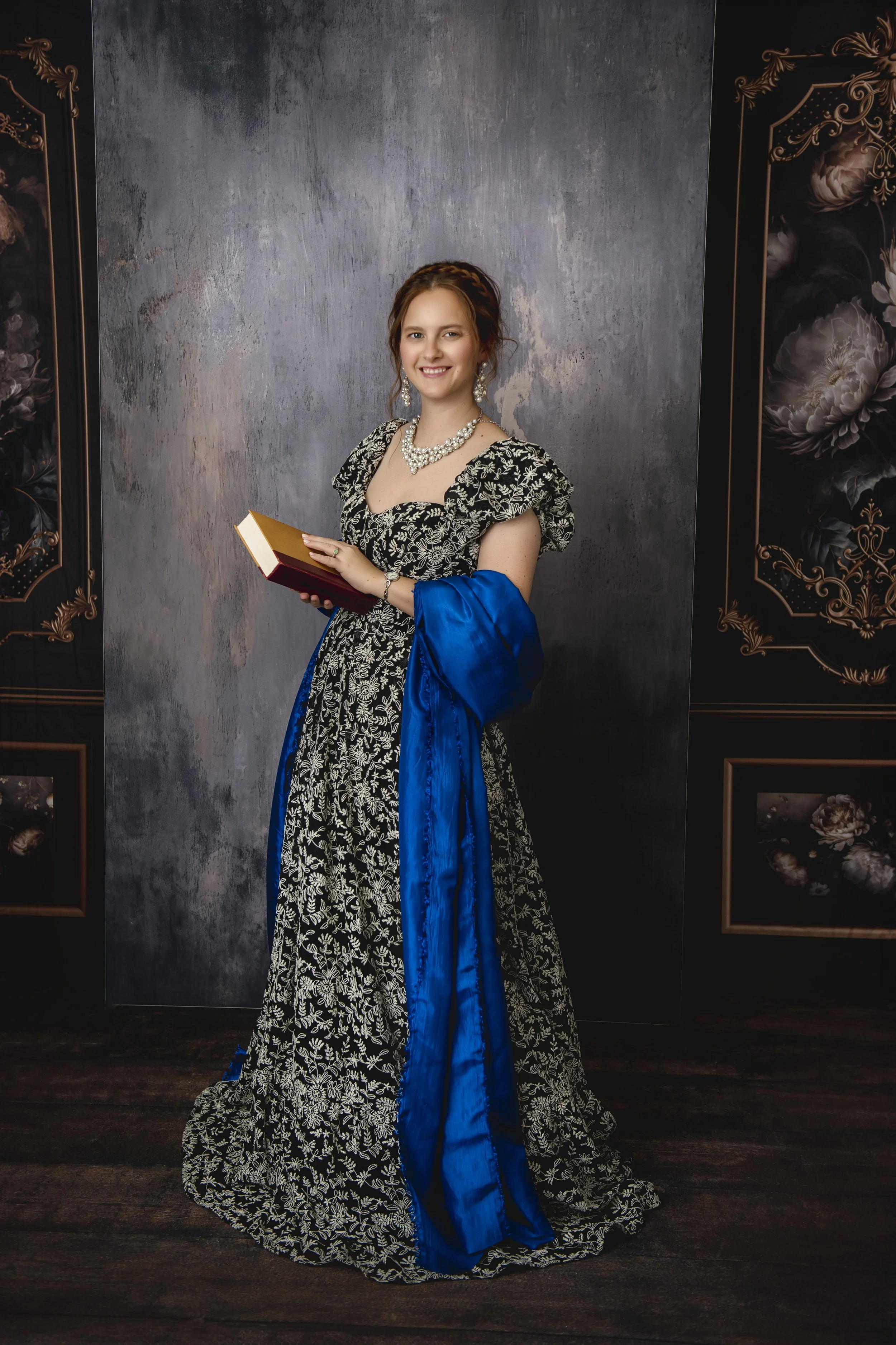 Young woman in a black and white floral gown with a bright blue sash, holding a book, standing in front of a painted background with dark floral designs.