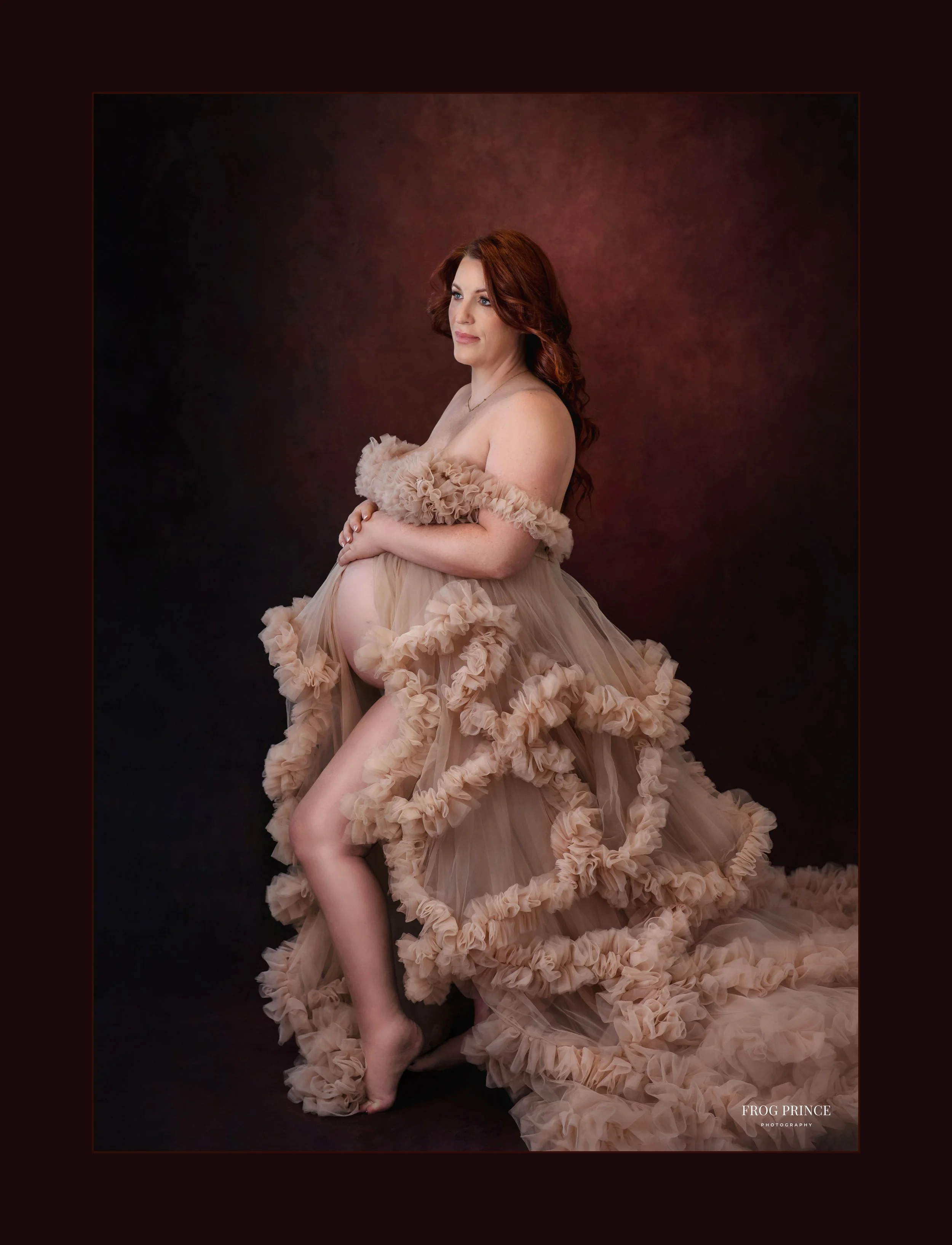 Heirloom maternity portrait of a pregnant mother with red hair wearing a ruffled beige gown, photographed against a dramatic dark backdrop in our luxury studio.