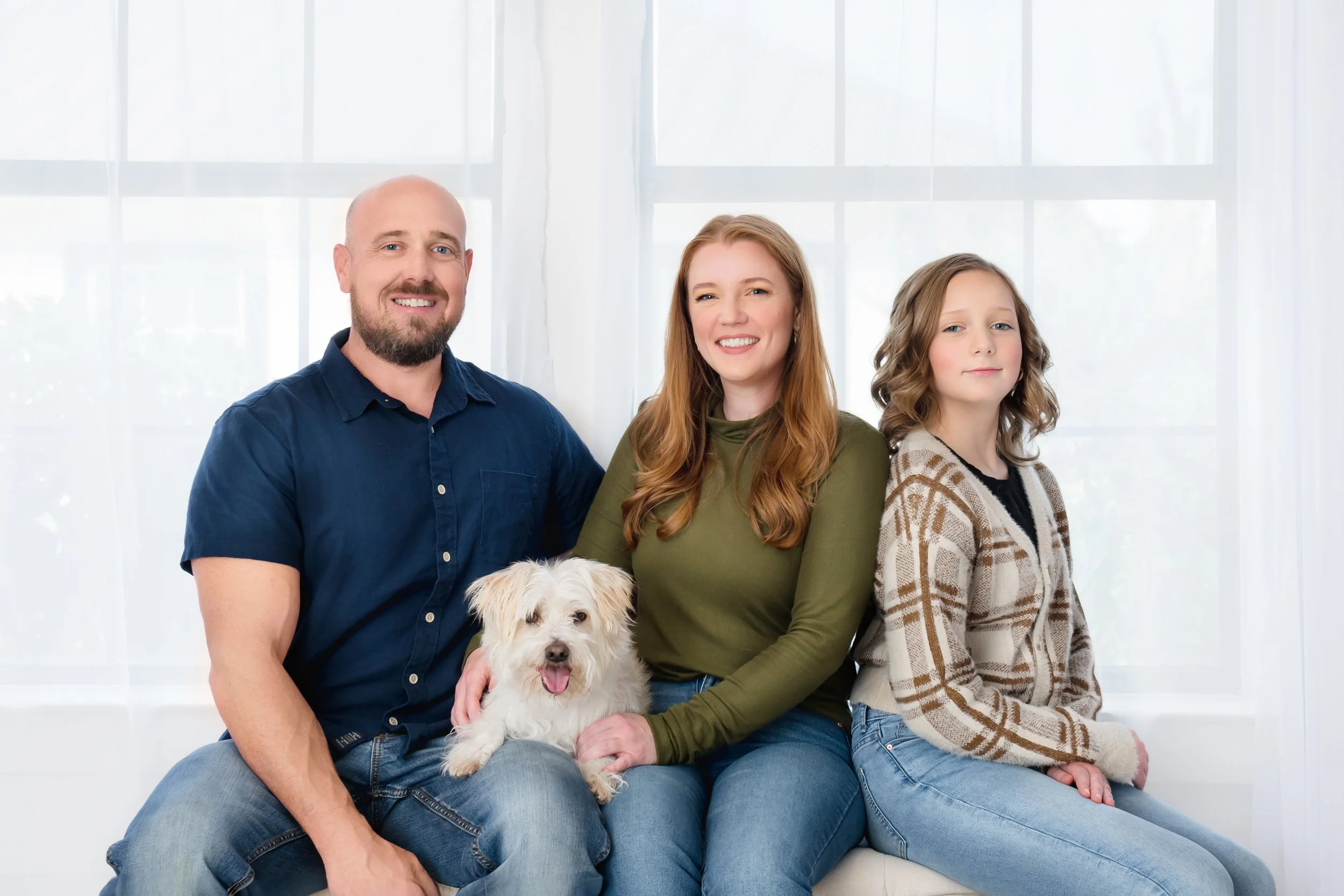 A family of three with a small white dog sitting on a white bench in front of a large window with sheer white curtains, smiling at the camera.