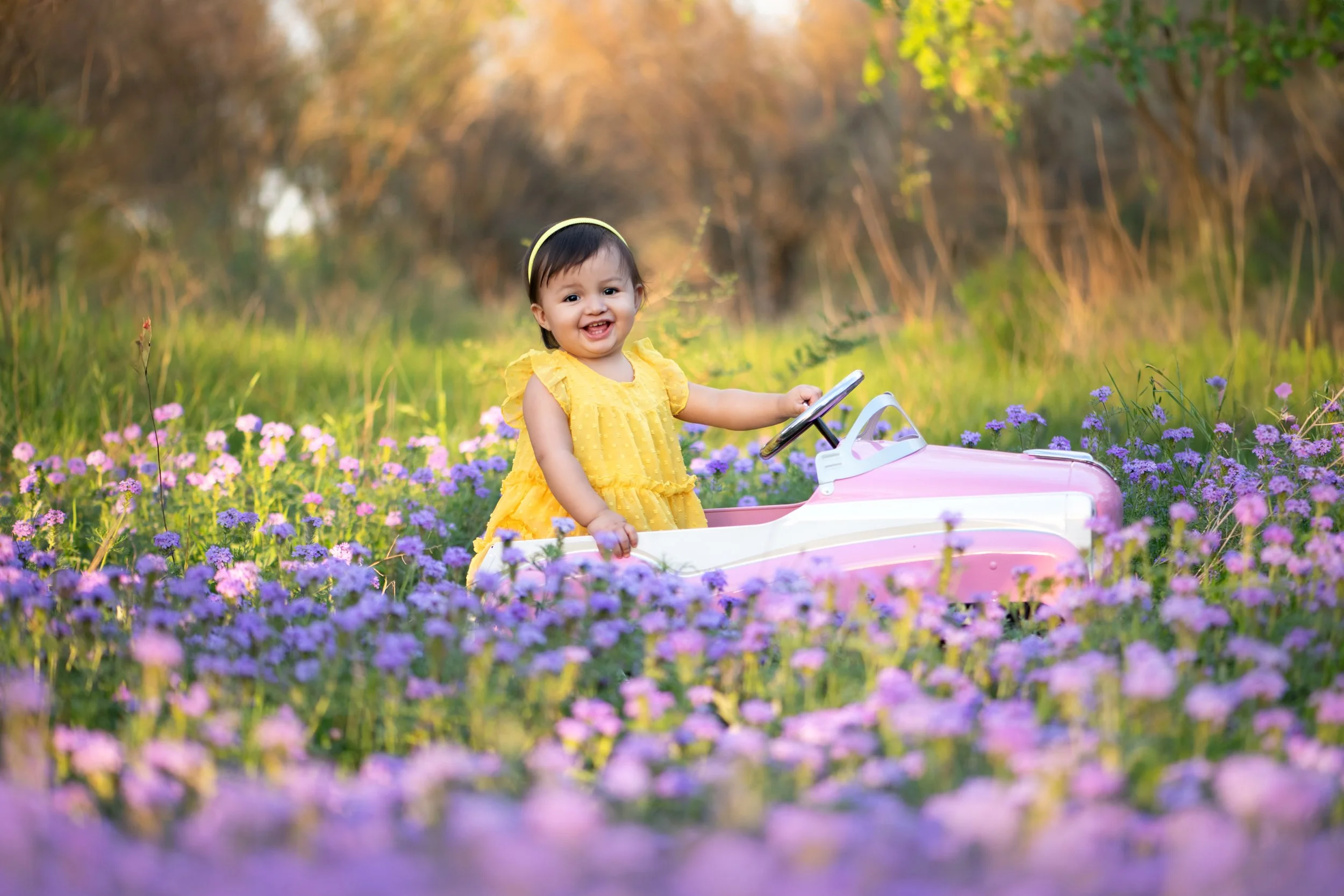 A young girl in a yellow dress sitting in a pink toy car in a field of purple flowers with trees in the background.