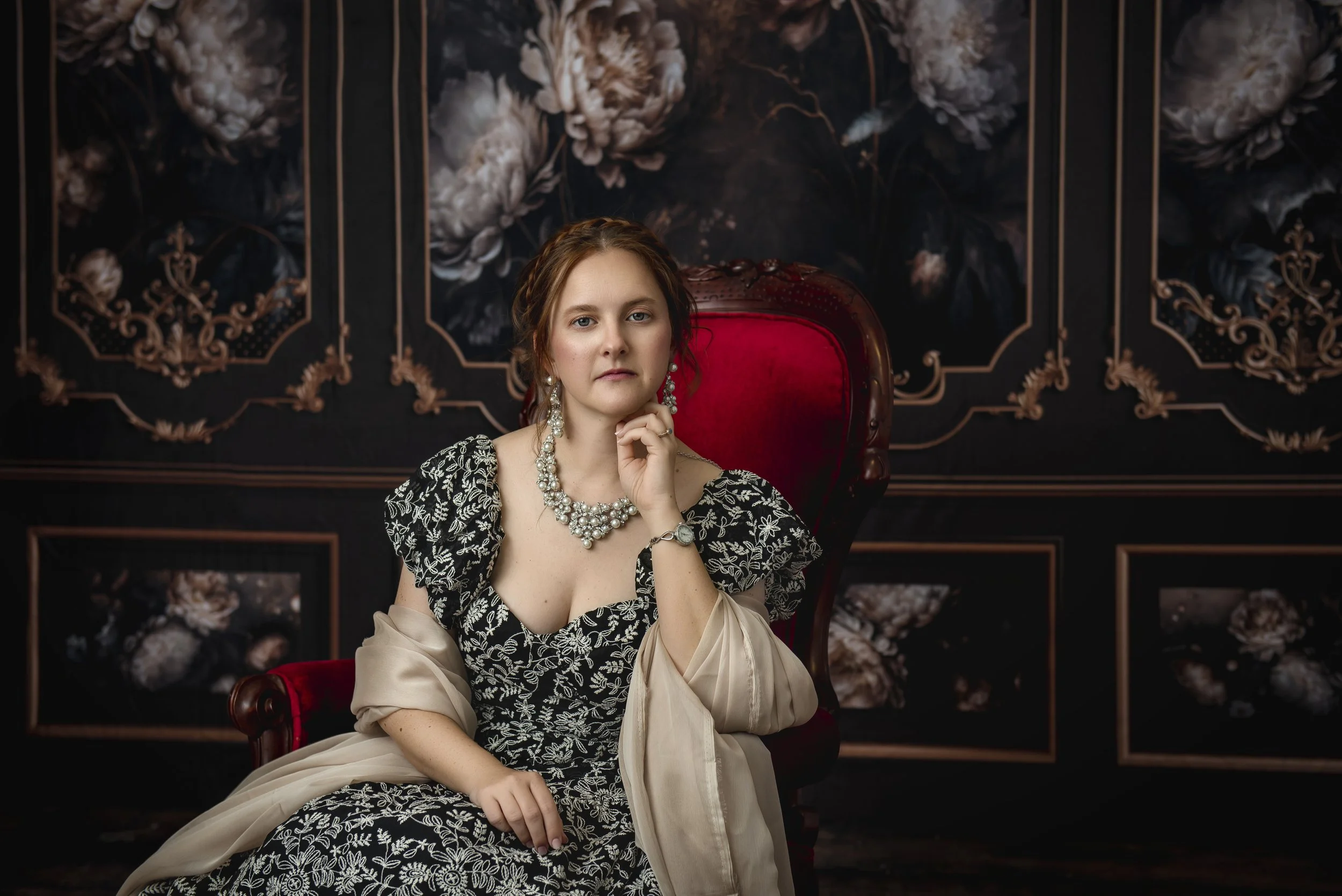 A woman with red hair and blue eyes sitting on a red velvet chair, wearing an elegant black and white floral dress, pearl jewelry, and a beige shawl, in a dark floral-patterned room.