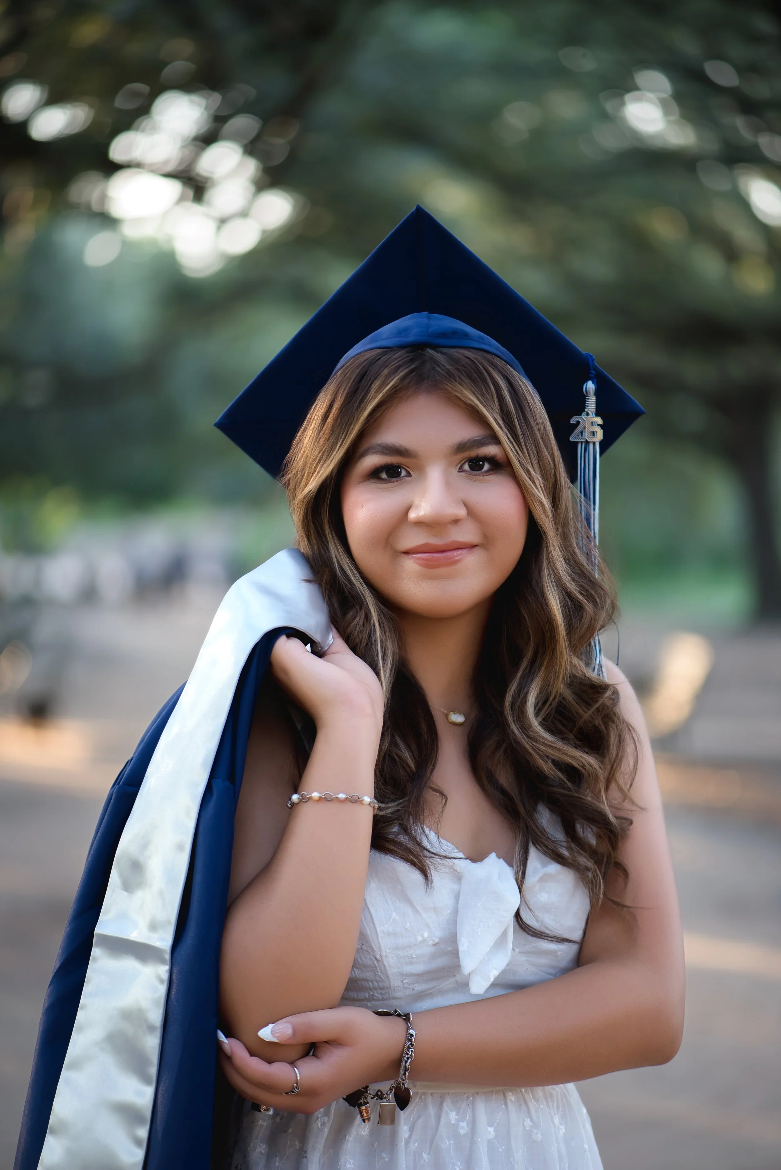 A young woman in a white dress and a blue graduation cap and gown, standing outdoors with trees in the background, smiling at the camera.
