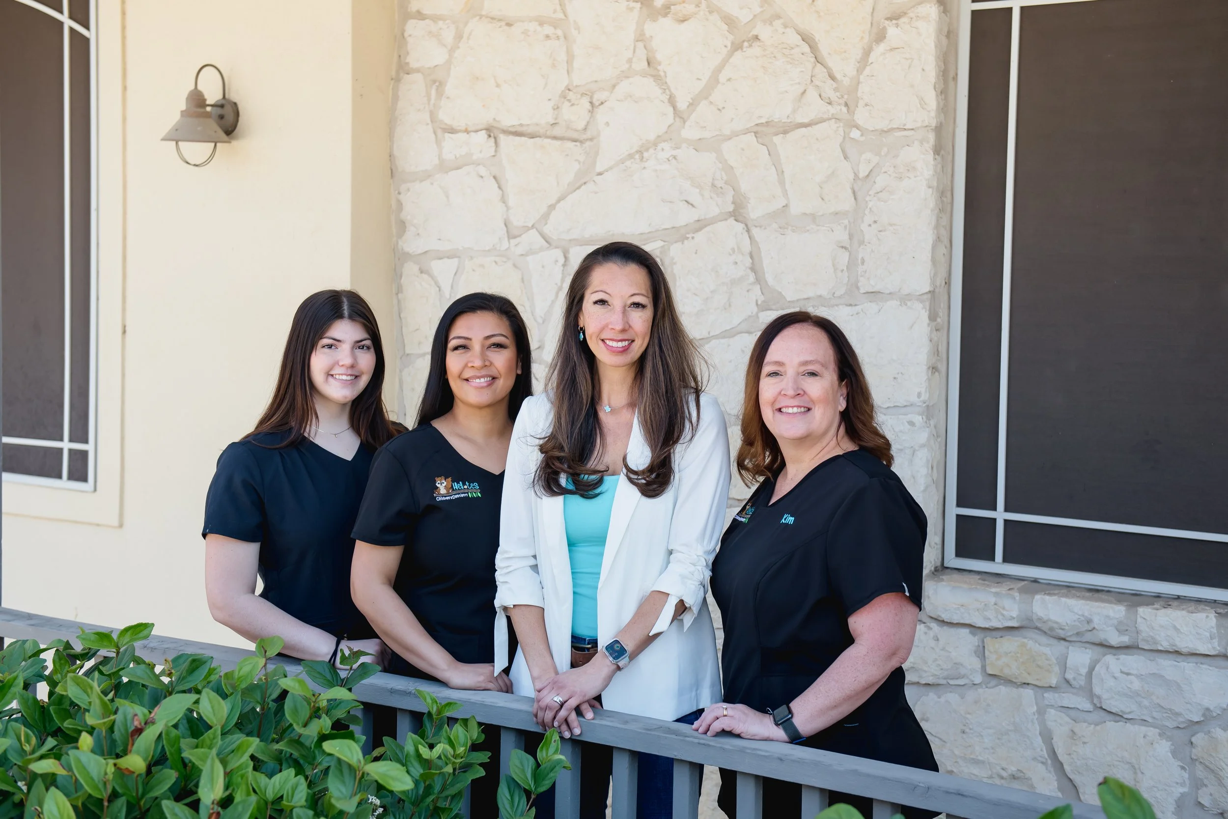 Four women standing outside near a stone wall and a window, smiling at the camera. One woman is in a white blazer, and the other three in black scrubs.