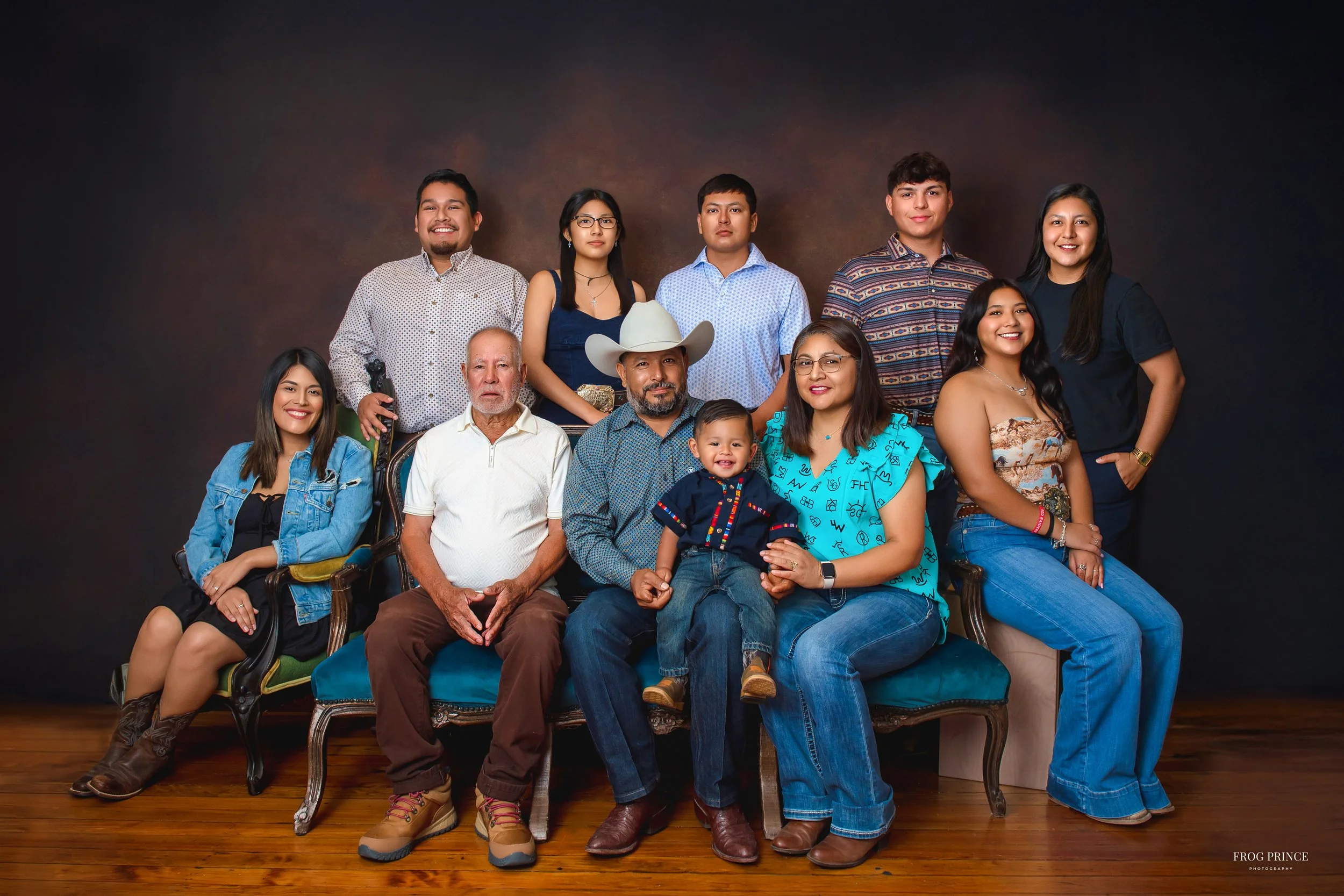 A multi-generational family photo with twelve members posing against a dark background, including children, teens, adults, and seniors, some sitting on chairs and others standing.
