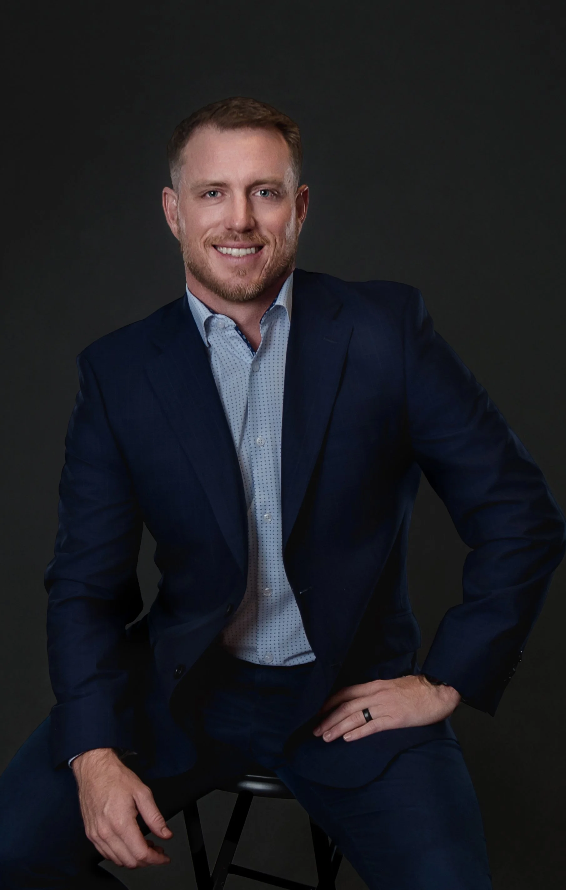 In our Spring Branch studio a man in a navy suit and light blue shirt sitting on a black stool against a dark background, smiling at the camera.