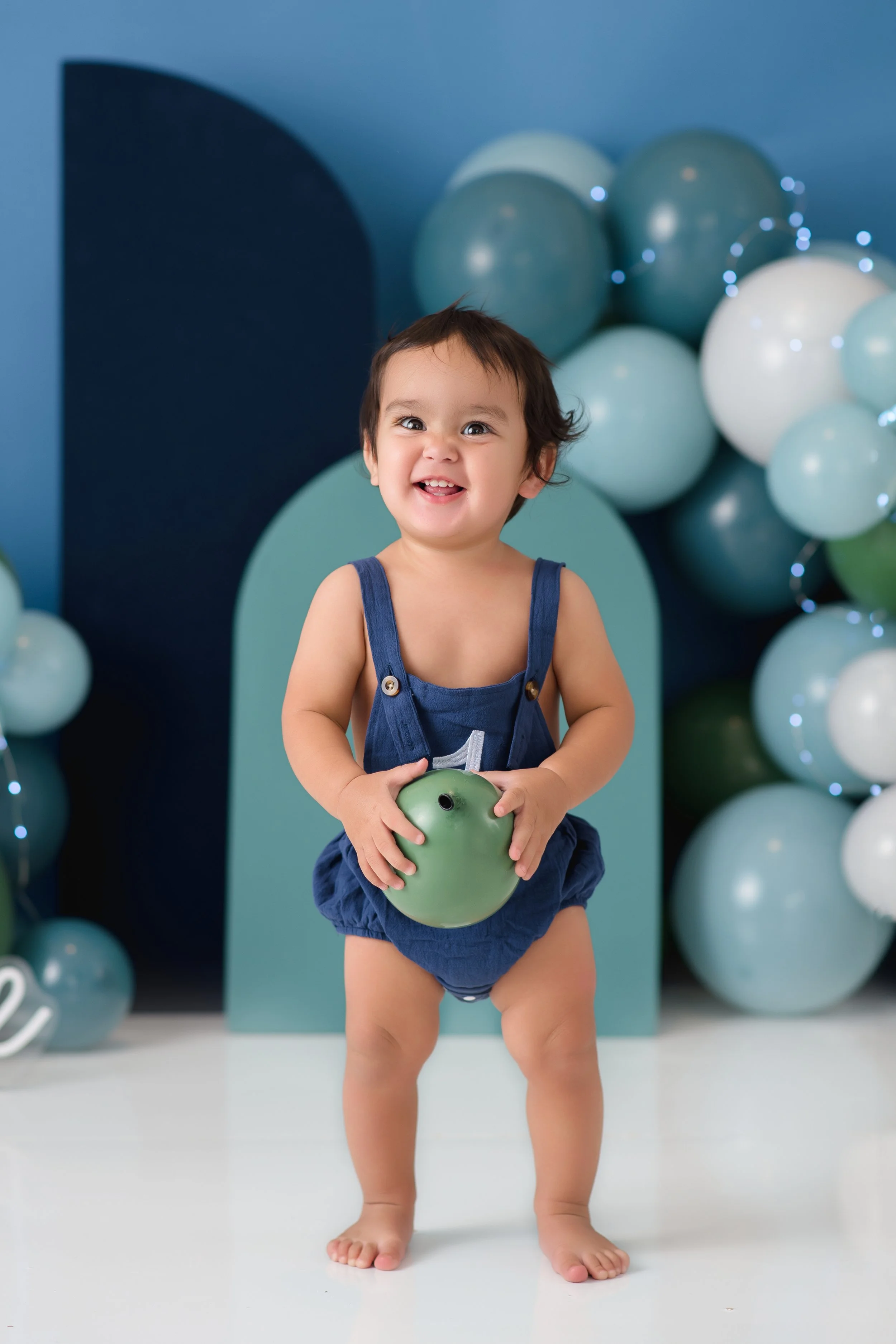 A smiling young child holding a green toy ball, standing barefoot in front of a blue background decorated with balloons and string lights.