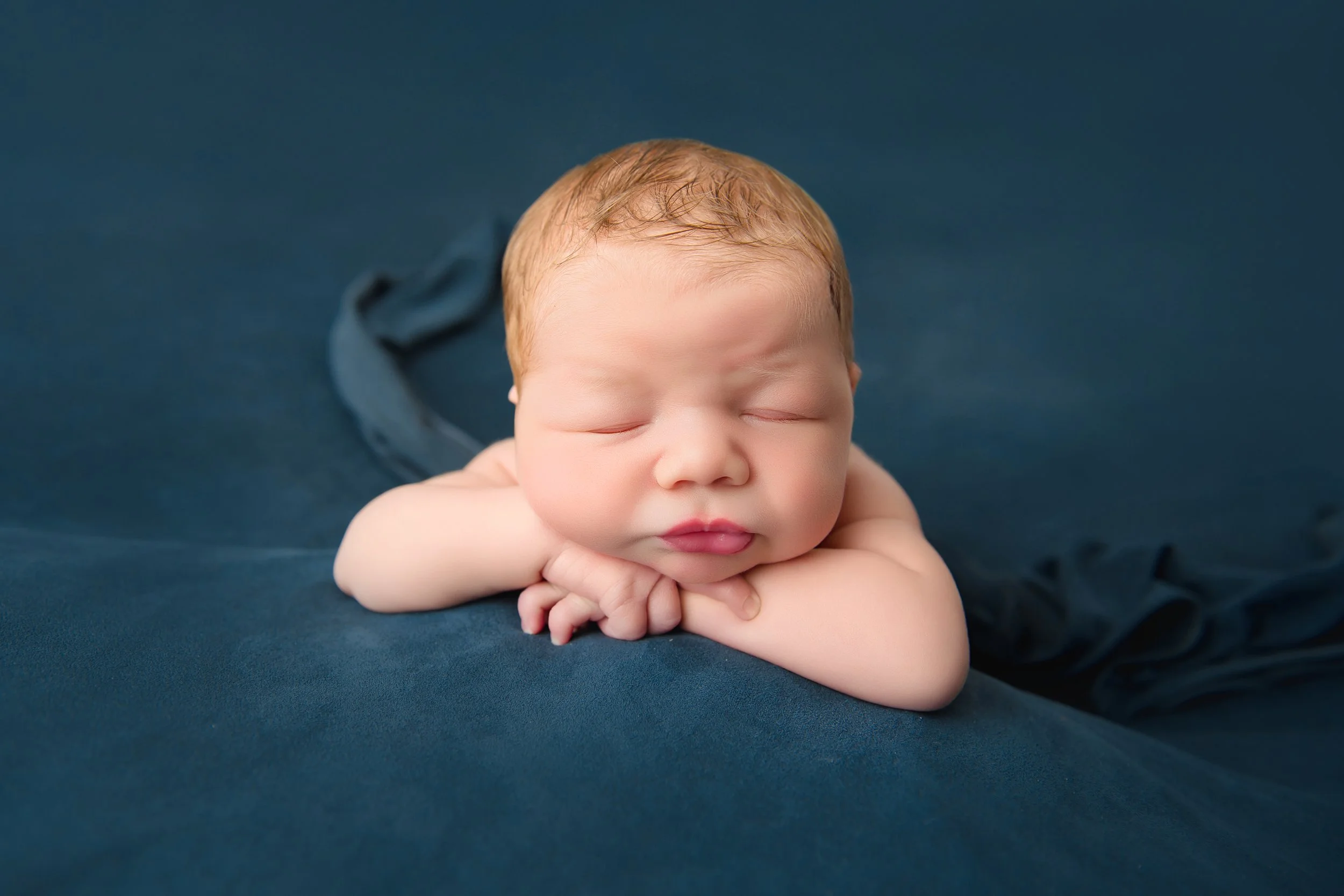 Close-up of a sleeping newborn baby with red hair, resting on a dark blue blanket, with eyes closed and arms folded under the chin.