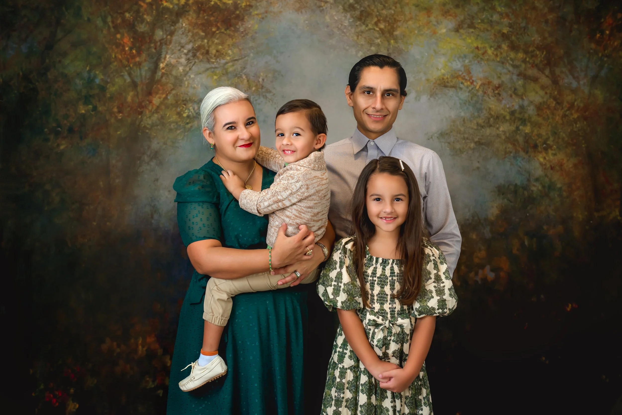 A family of five posing for a portrait outdoors with a blurred autumnal background. The grandmother has gray hair and wears a green dress, holding her young grandson. The grandfather is dressed in a light gray shirt, standing behind their granddaught