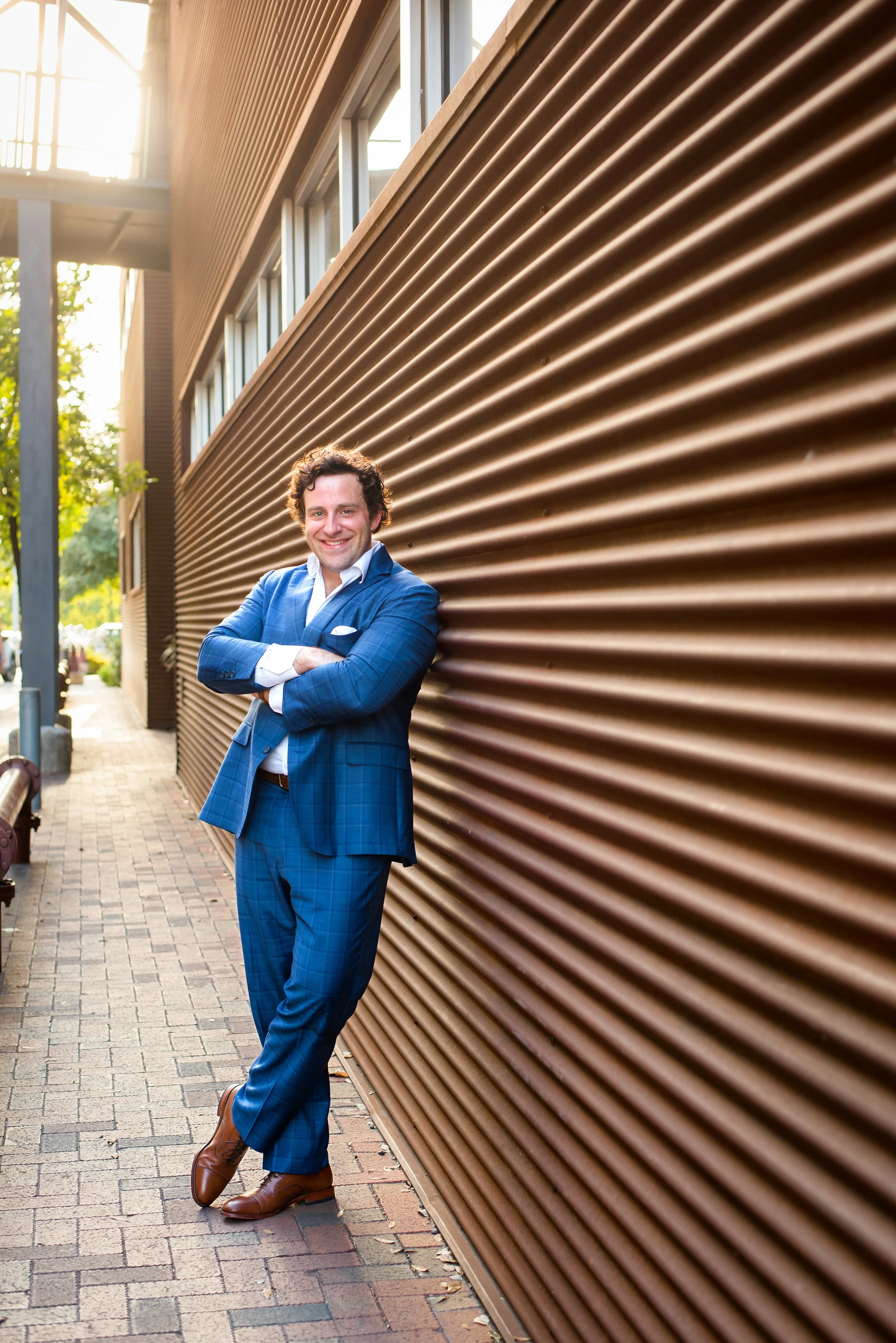 A smiling man in a blue plaid suit stands with arms crossed against a metallic brown corrugated wall on a city sidewalk during daylight.