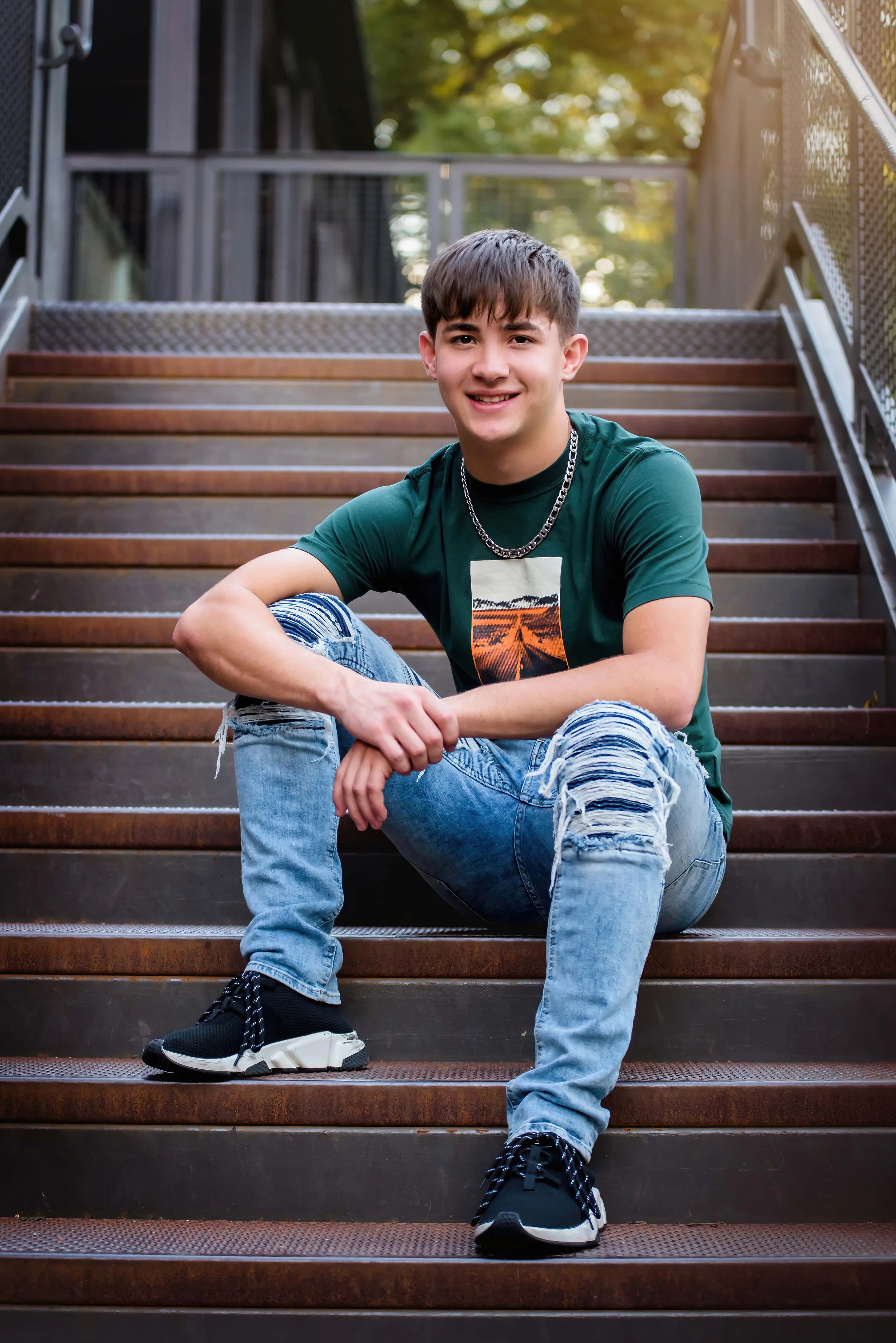 A young man with short brown hair and a bright smile sits on outdoor metal stairs, wearing a green graphic t-shirt, distressed jeans, sneakers, and a chain necklace.