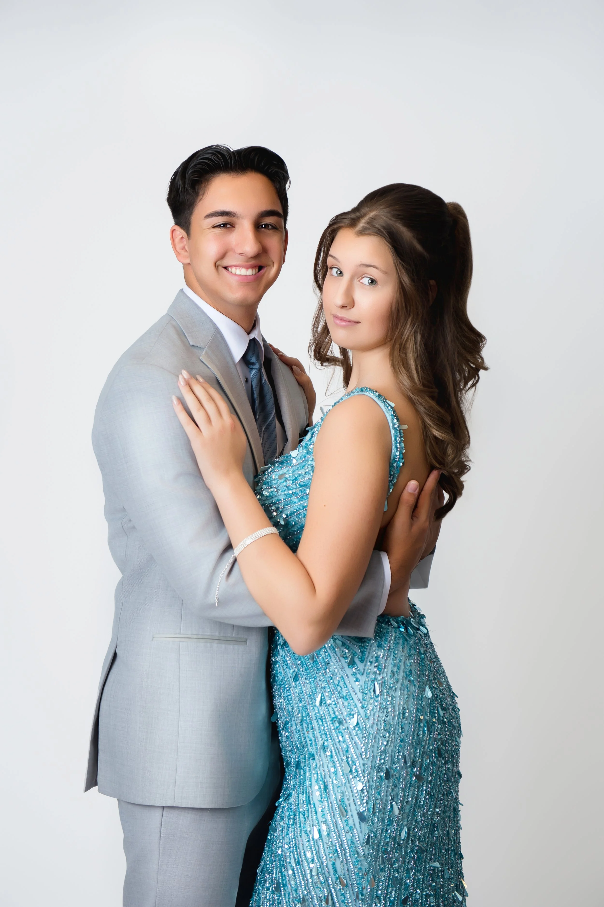 A young couple in formal attire, standing closely together against a plain white background. The man is in a light gray suit, and the woman is wearing a sparkling, blue, floor-length gown in our Spring Branch studio.