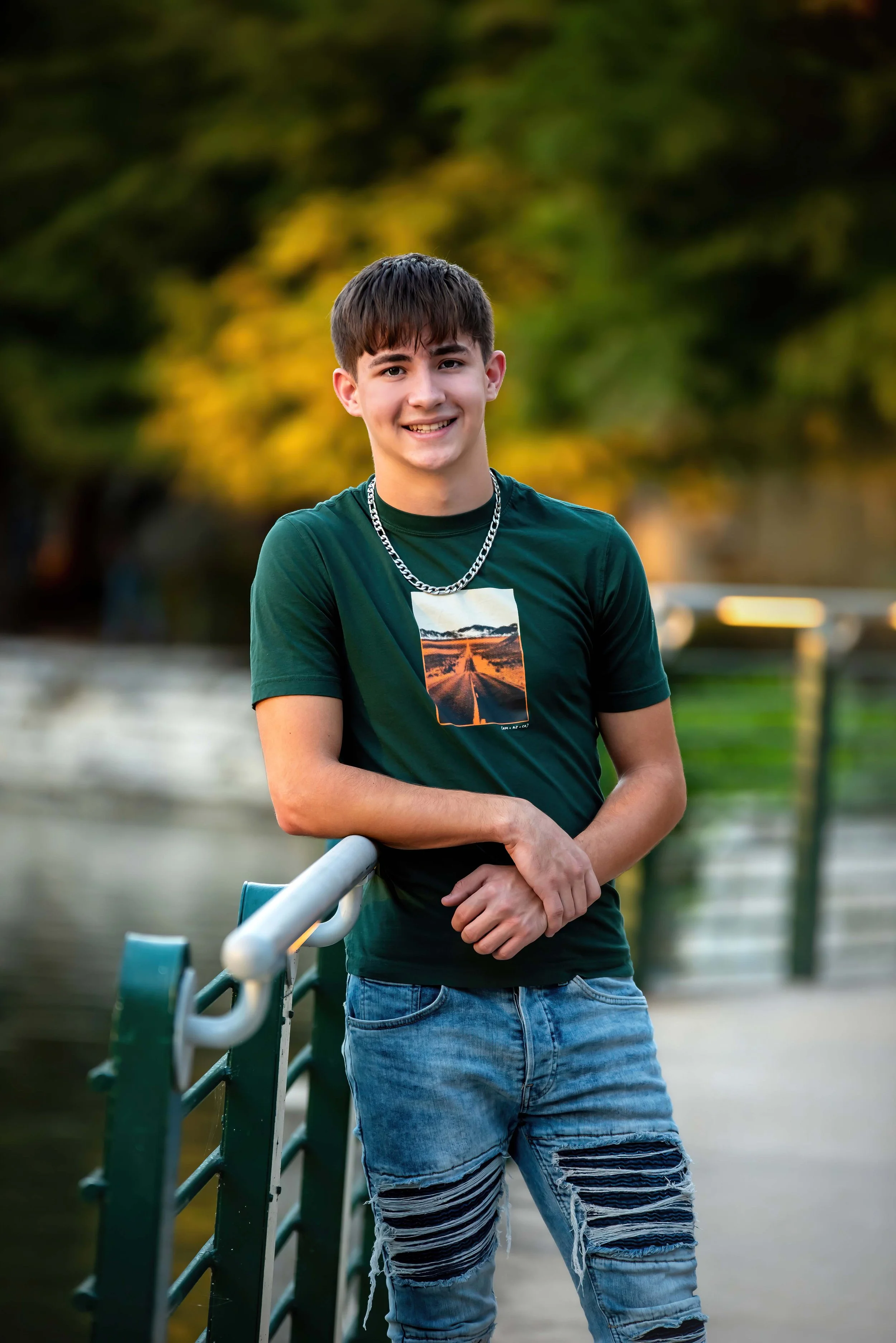 A young man with short dark hair smiling outdoors, wearing a green graphic t-shirt, a silver chain necklace, and ripped jeans, standing by a railing near water with trees in the background.
