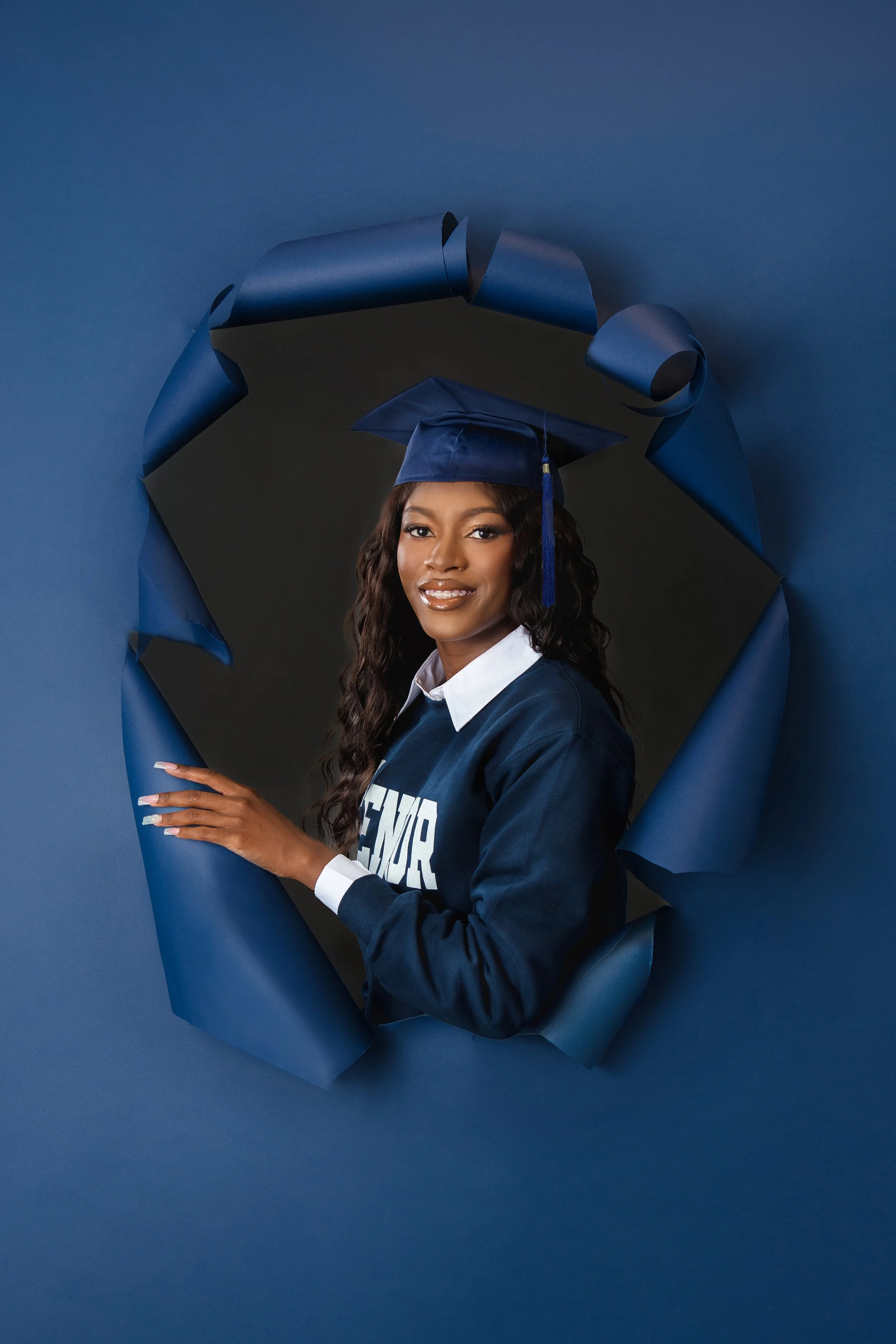 In our Spring Branch studio a young woman wearing a graduation cap and gown, standing behind a torn blue paper backdrop, smiling at the camera.