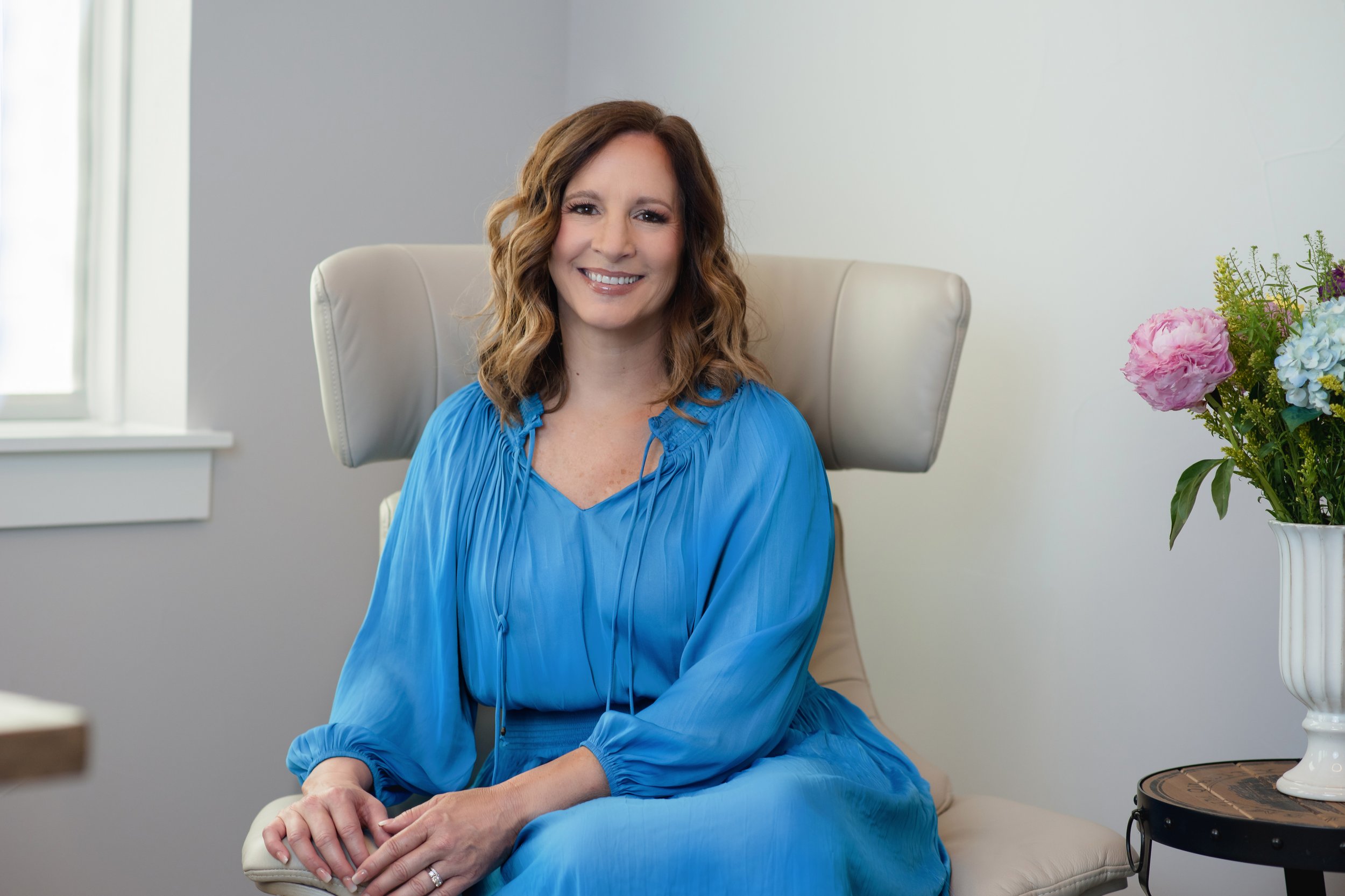 A woman with shoulder-length wavy brown hair wearing a bright blue dress, sitting on a cream-colored chair, smiling in a well-lit room with a window to her left and a bouquet of colorful flowers in a white vase on a small table to her right.