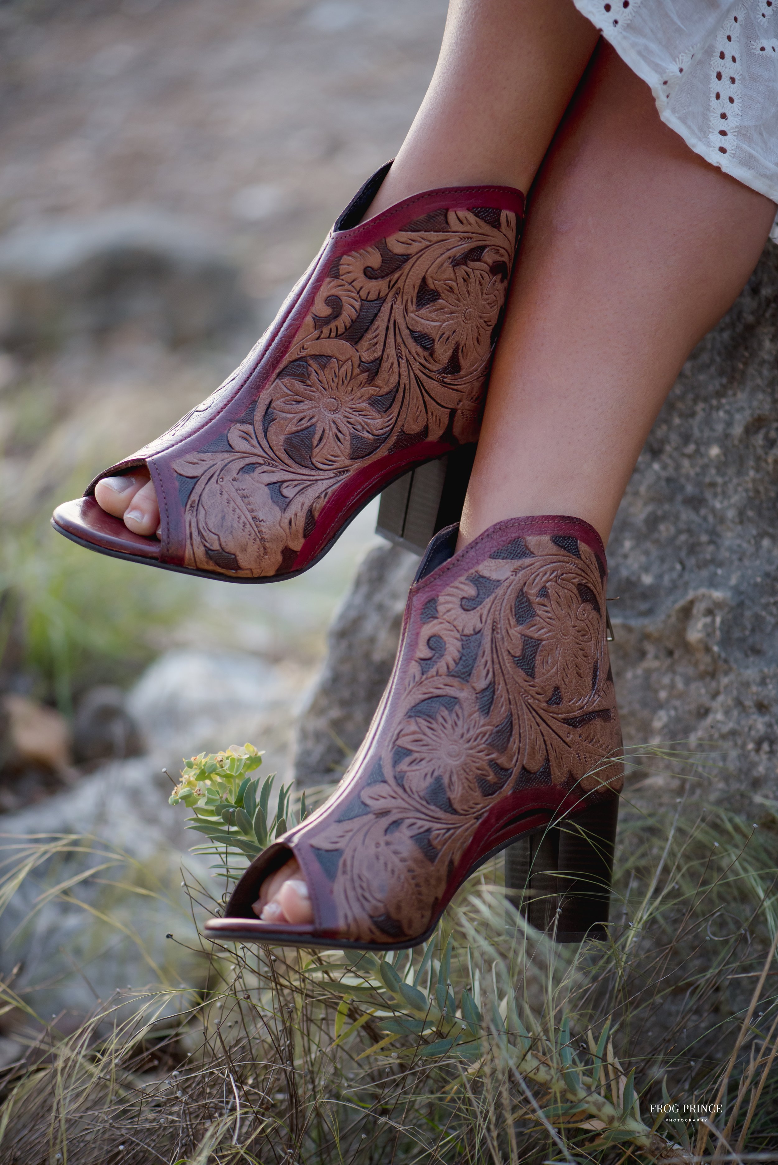 Close-up of floral pattern leather open-toe high-heeled boots worn by person sitting on a rock outdoors with grass and rocks visible in the background.