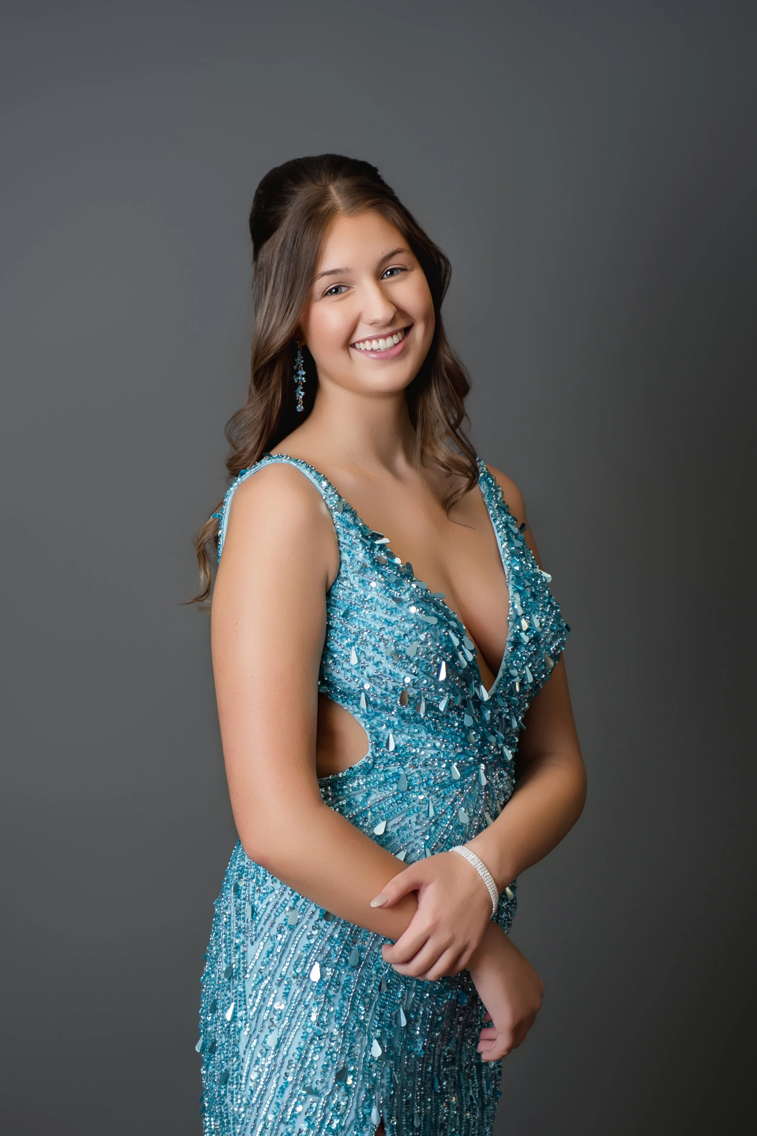 Young woman in a glamorous turquoise beaded evening gown with sequins, posing against a plain gray background, smiling in our Spring Branch studio.