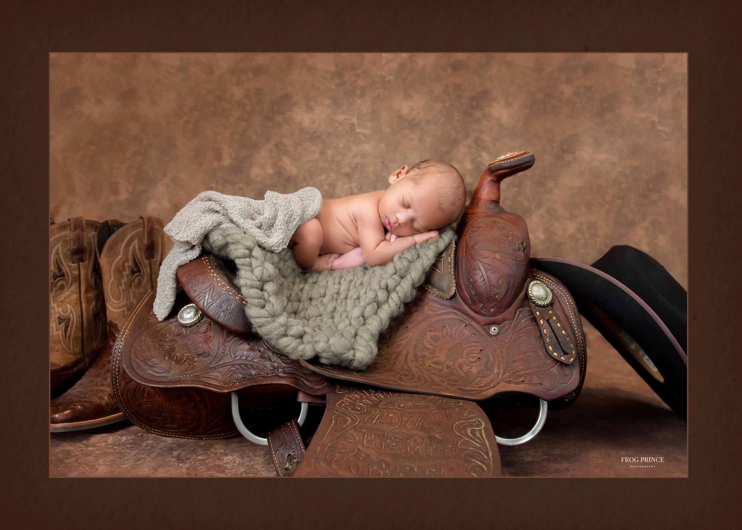 A newborn baby sleeping curled up on a decorative Western saddle with a knitted blanket and a soft gray cloth, brown background.