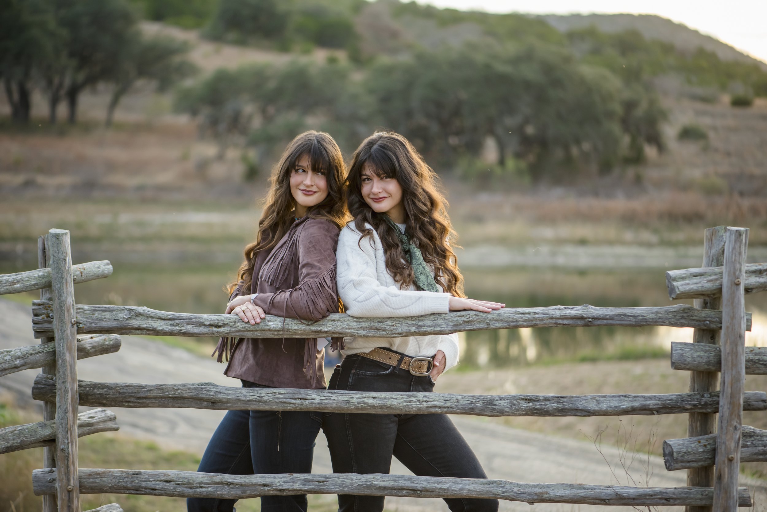 Two women with long, wavy hair standing outdoors behind a wooden fence near a body of water with trees and hills in the background.
