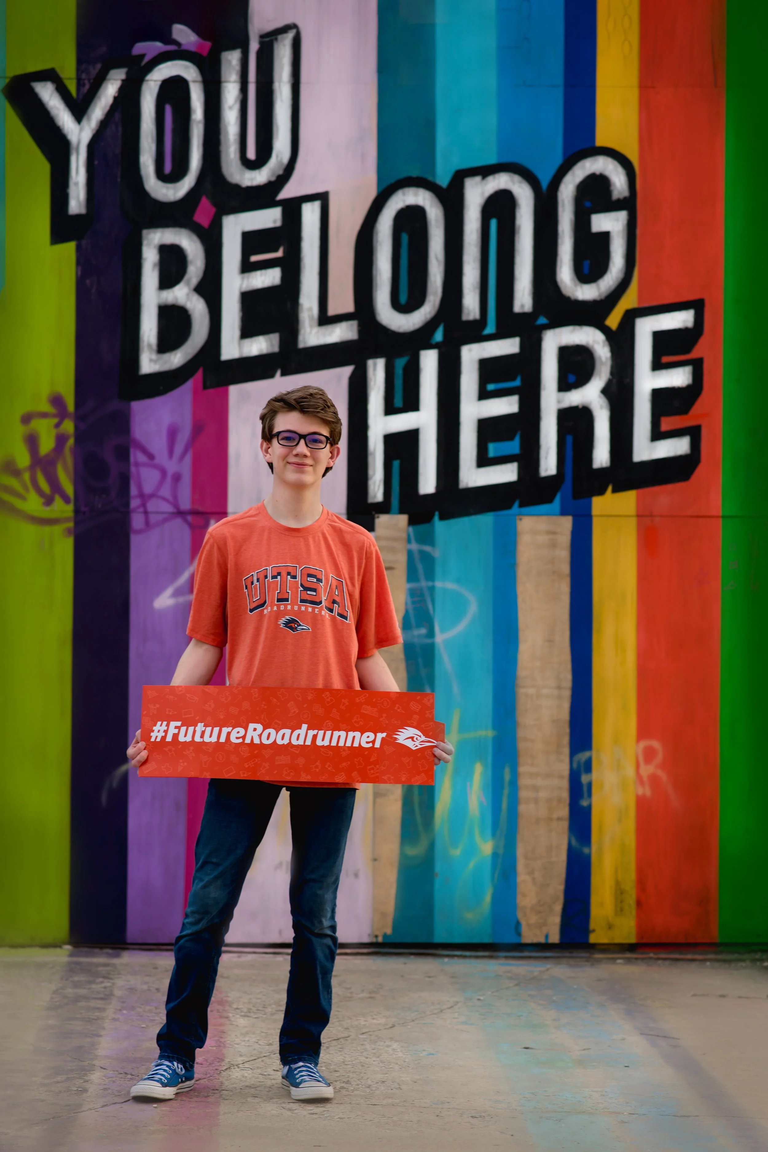 A young man with glasses wearing an orange UTSA t-shirt stands in front of a colorful mural that reads 'You Belong Here.' He holds a red sign that says '#FutureRoadrunner' with the UTSA logo.