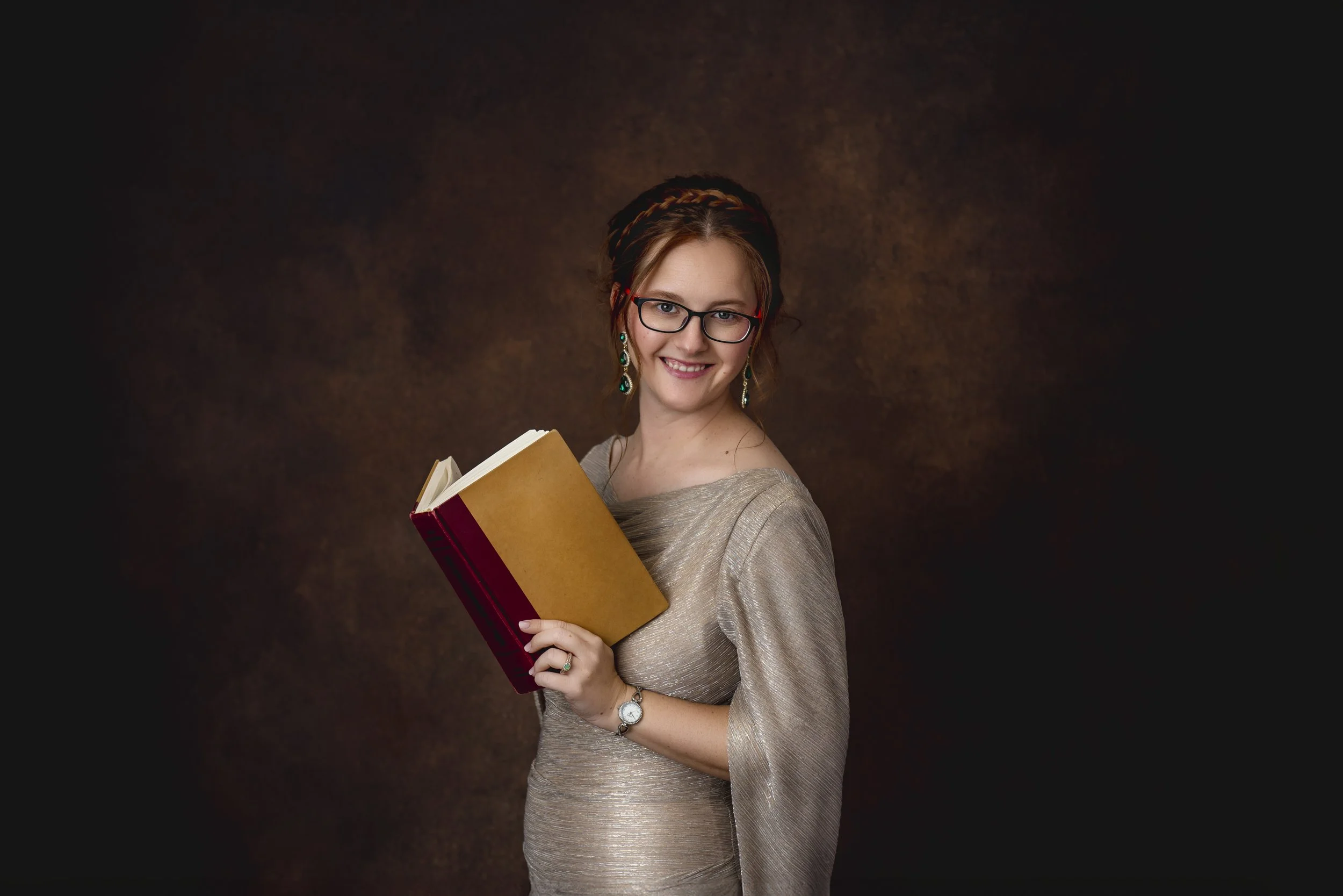 A young woman wearing glasses and earrings, holding an open book and smiling, standing against a dark brown textured background in our Spring Branch studio.
