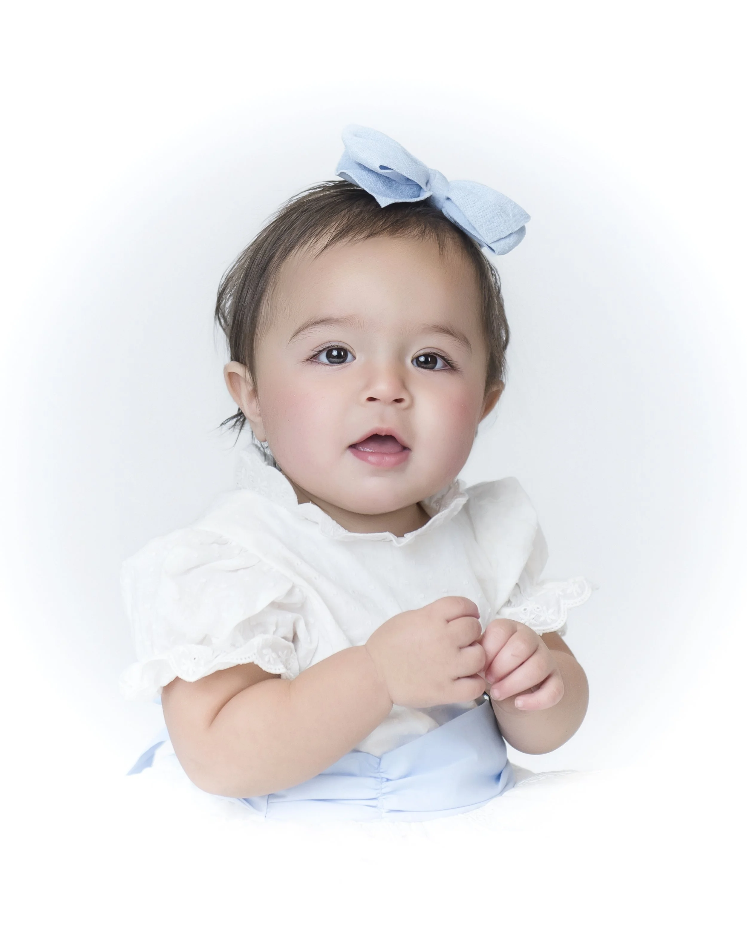 A young child with brown hair, wearing a white dress with lace details and a blue bow on her head, looks at the camera with a neutral expression, set against a plain white background.