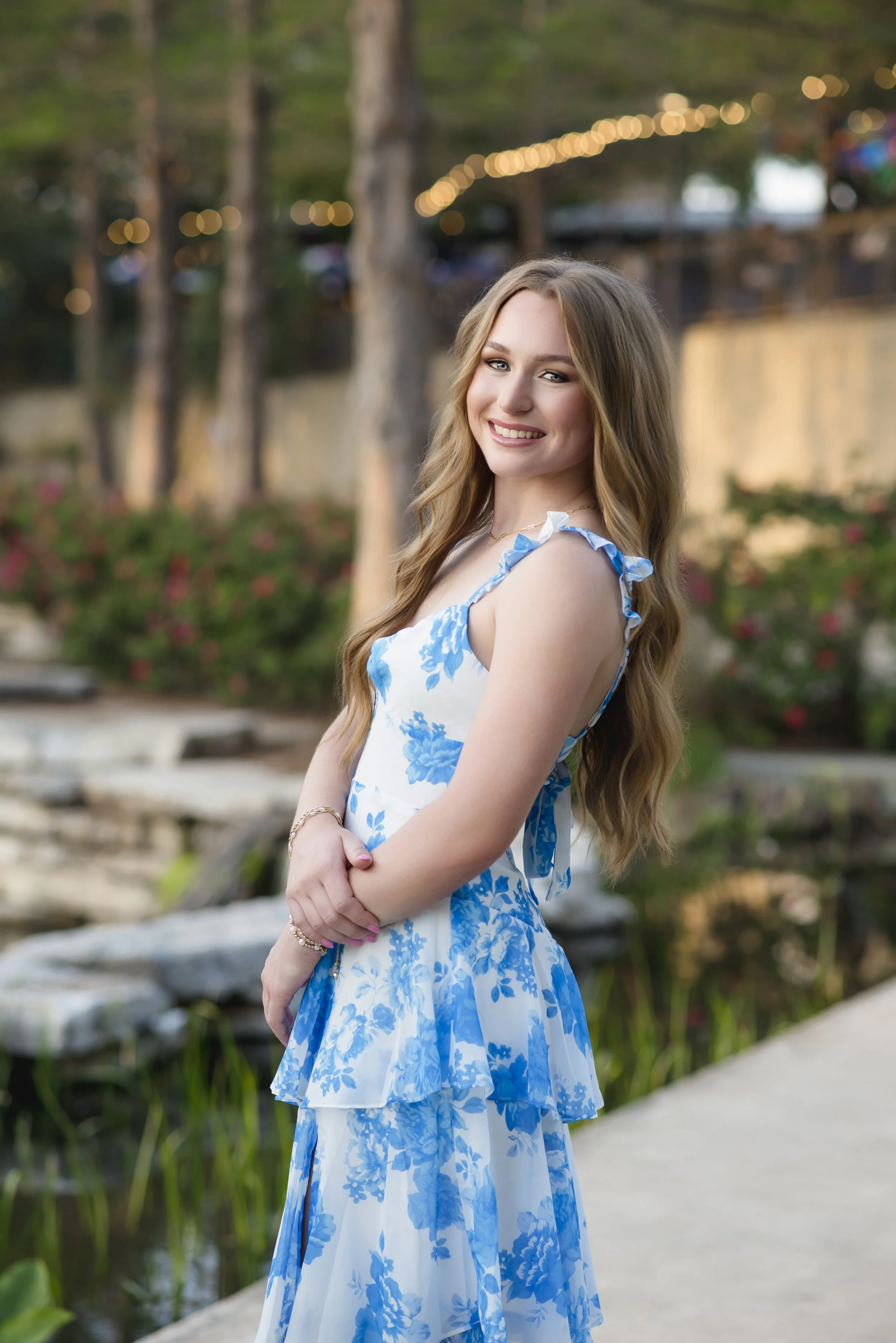 A young woman with long, wavy light brown hair smiling at the camera, wearing a blue and white floral dress, standing outdoors near a pond with greenery, trees, and string lights in the background during the evening.
