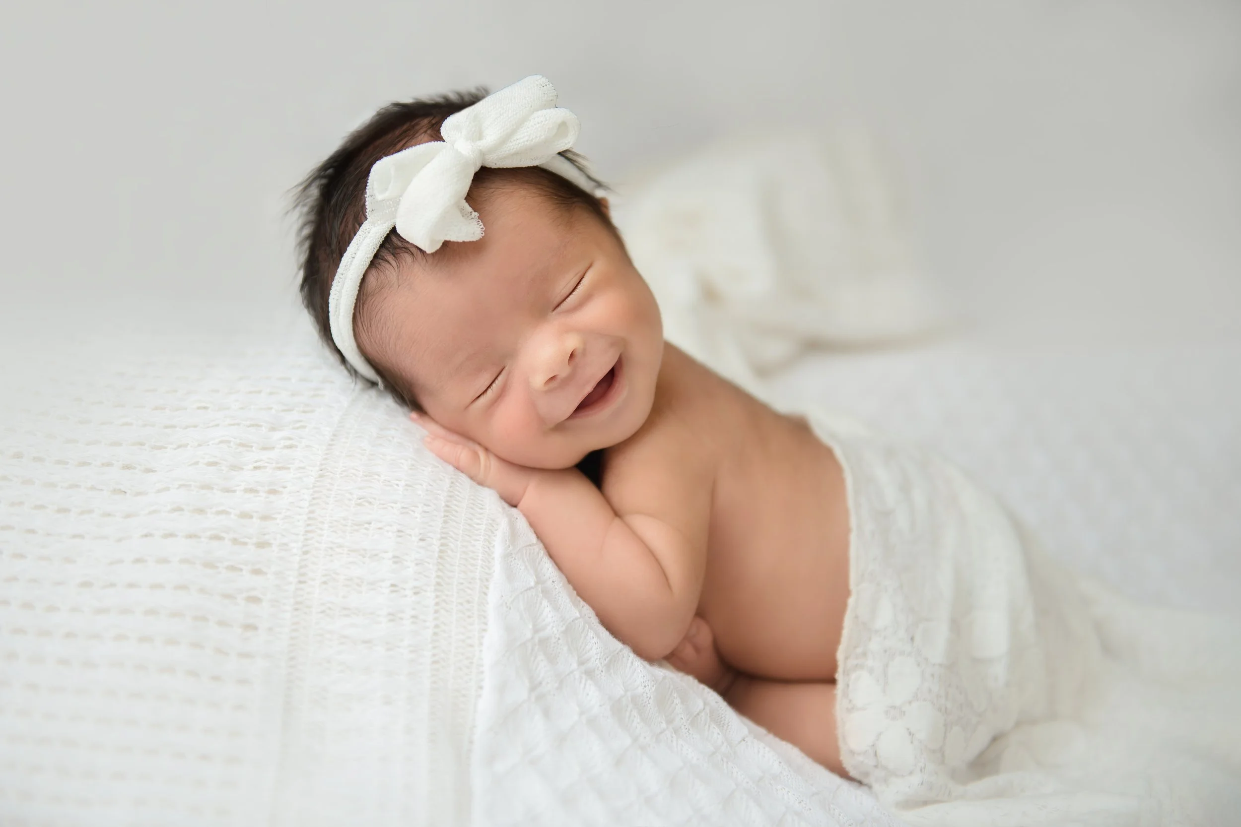 Light and airy newborn portrait of a smiling baby wearing a white bow headband, posed on a soft textured white blanket.