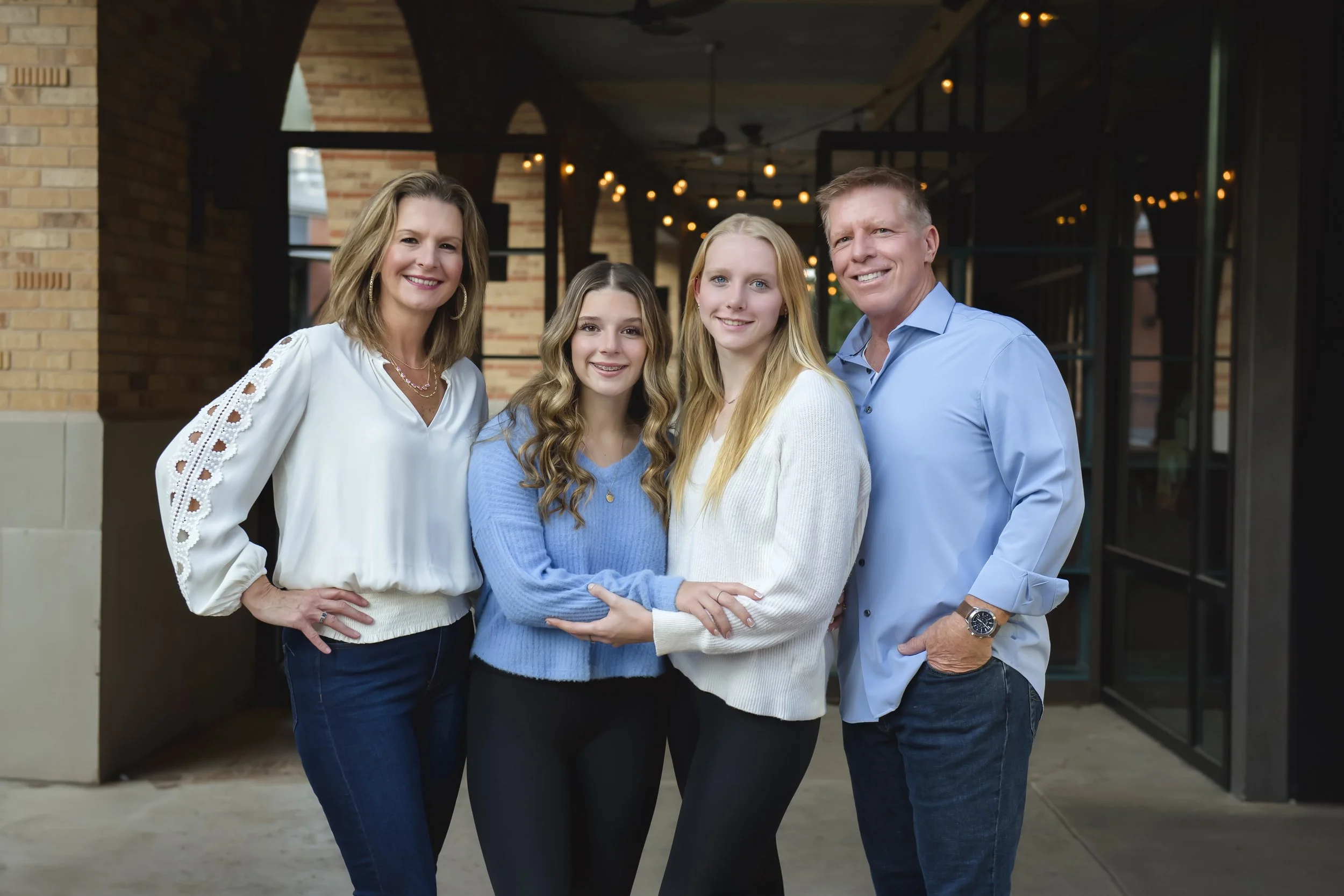 A family of four standing close together outdoors, smiling at the camera. The woman on the left is wearing a white blouse and jeans, the girl next to her has long wavy hair and is wearing a blue sweater, the girl next to her has long straight hair an