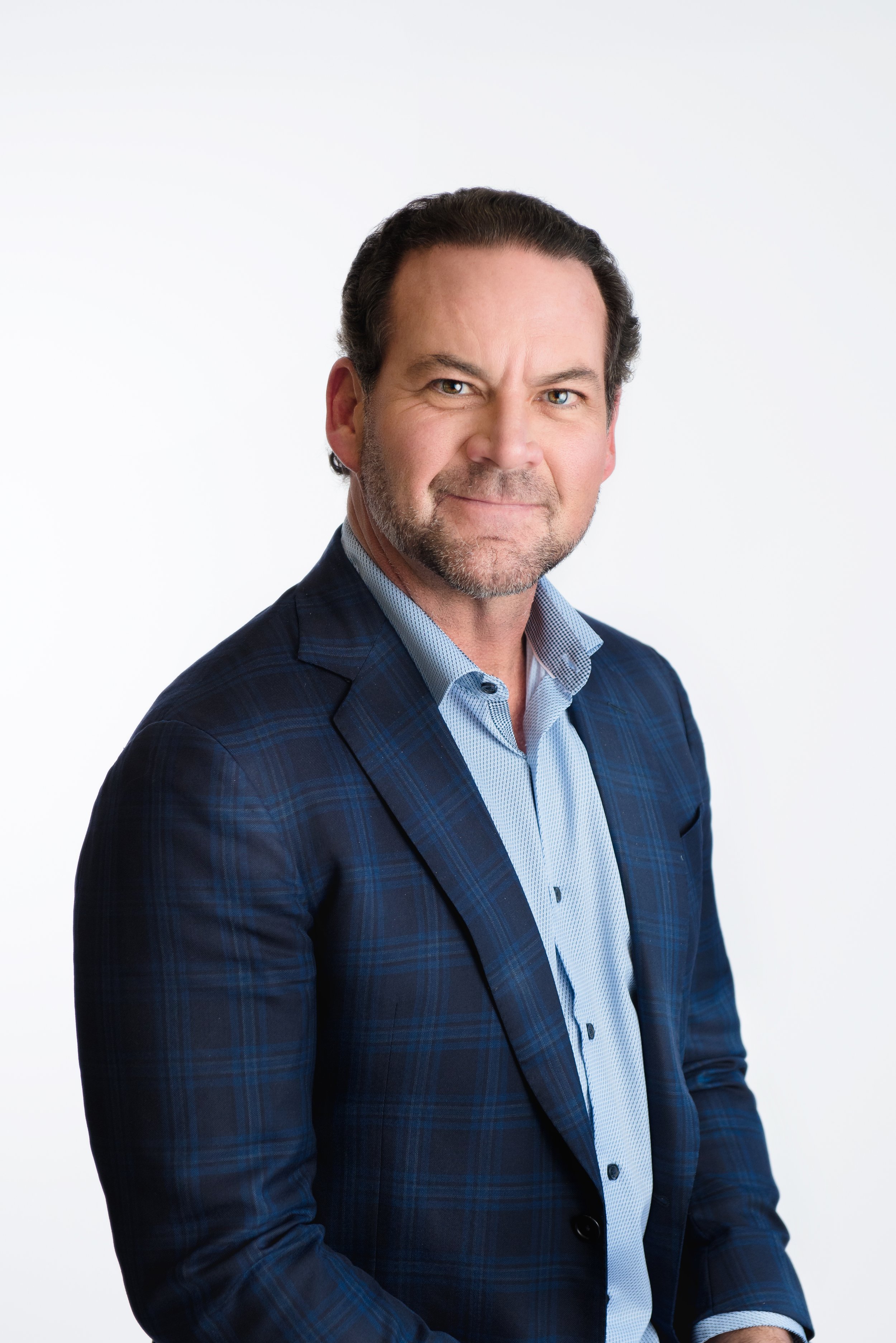 In our Spring Branch studio a professional man wearing a dark blue plaid blazer and a light blue dress shirt, standing against a plain white background.