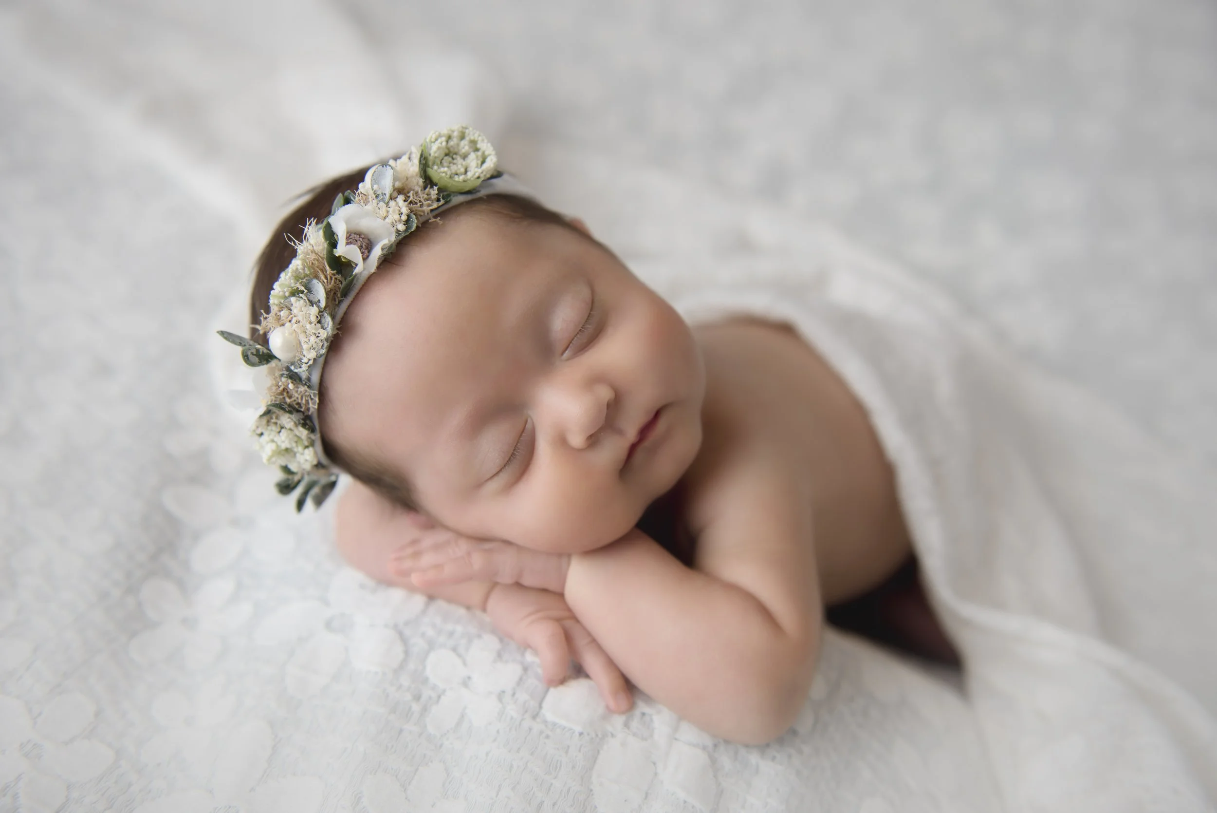 A sleeping baby with a floral headband, resting on a white textured blanket.