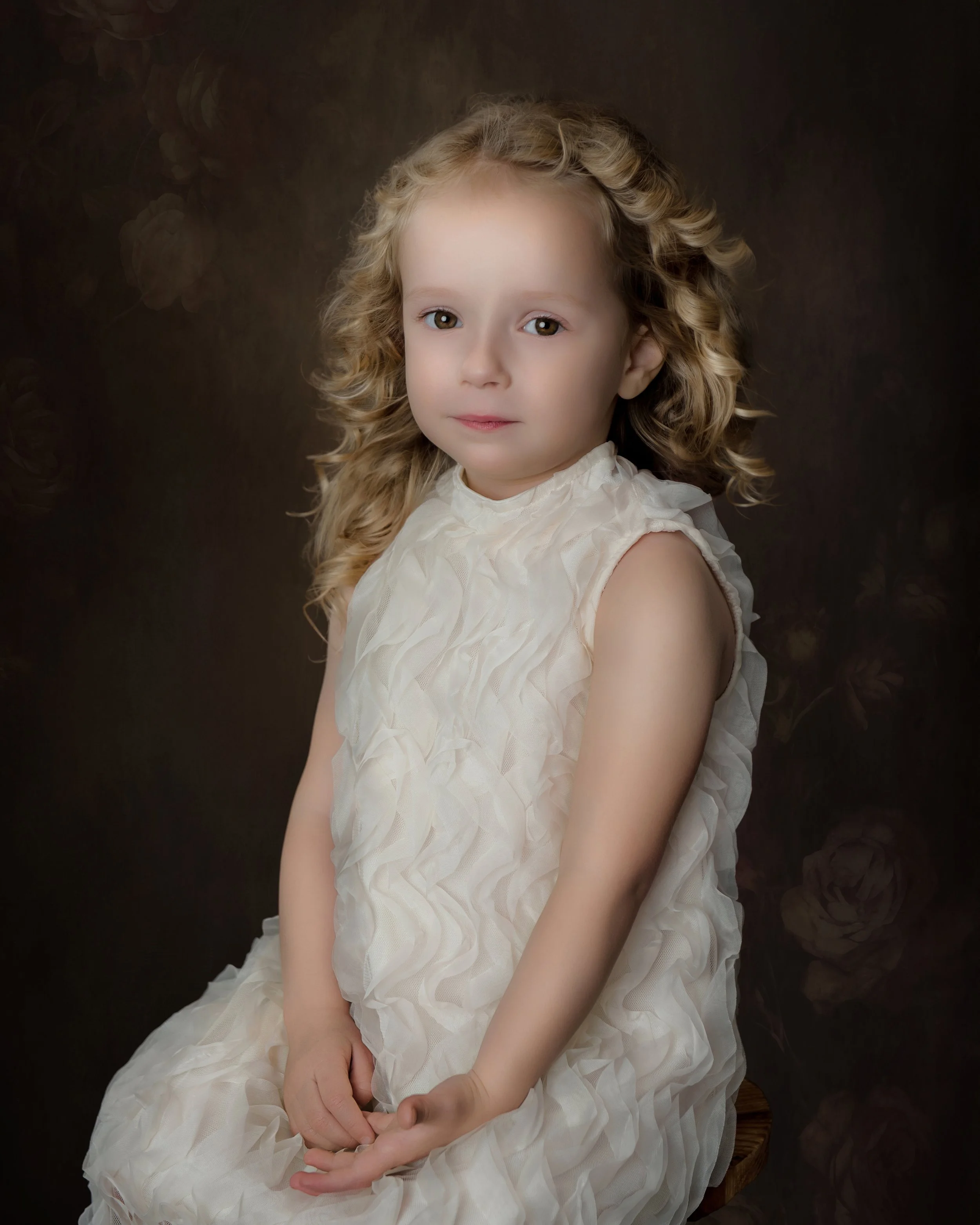 A young girl with curly blonde hair wearing a cream-colored, ruffled dress, sitting against a dark background.