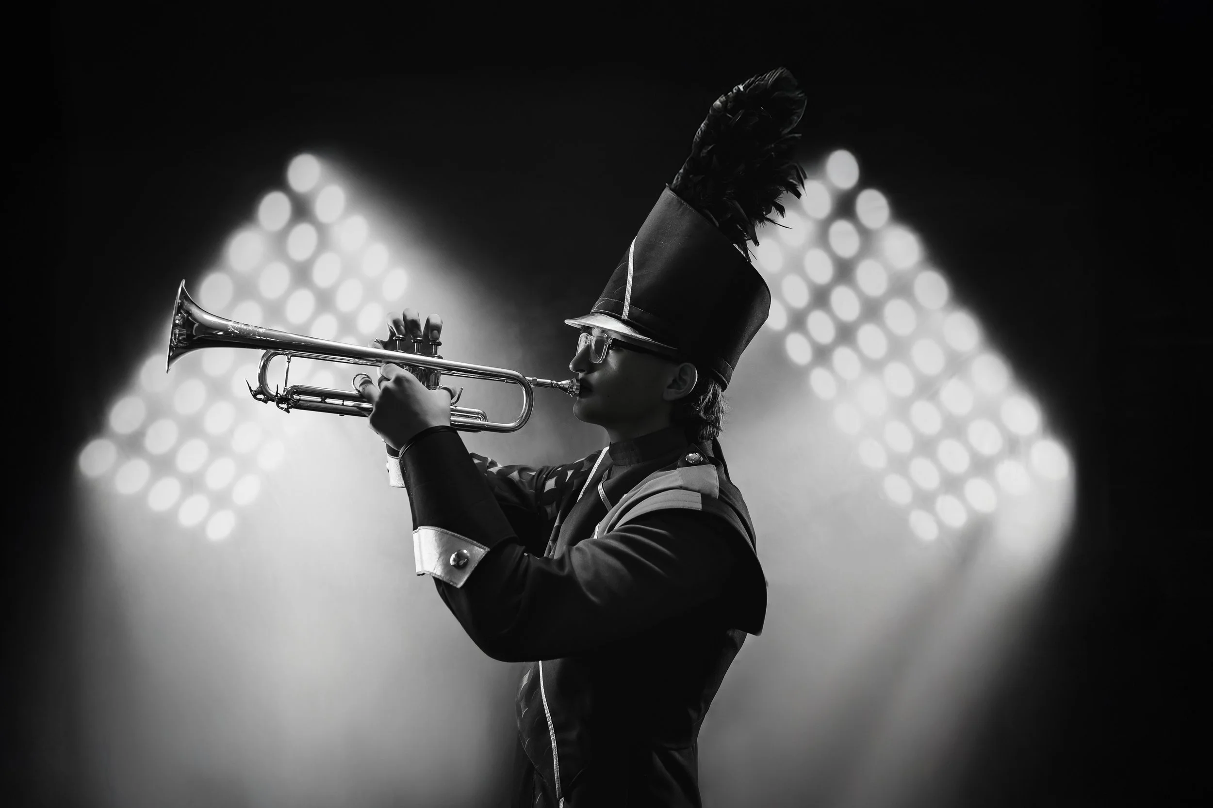 A person dressed in a marching band costume and tall hat with a feather, playing a trumpet on stage with bright lights in the background.