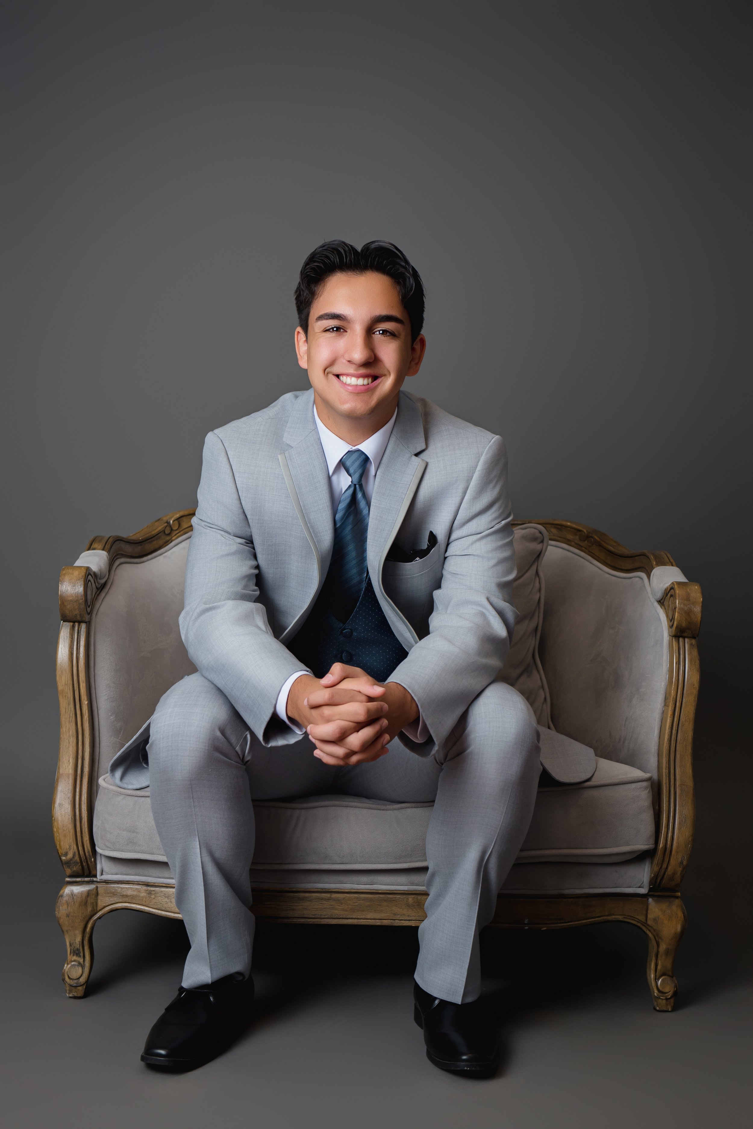 Young man in a light gray suit sitting on an ornate vintage couch, smiling, with a plain gray background.