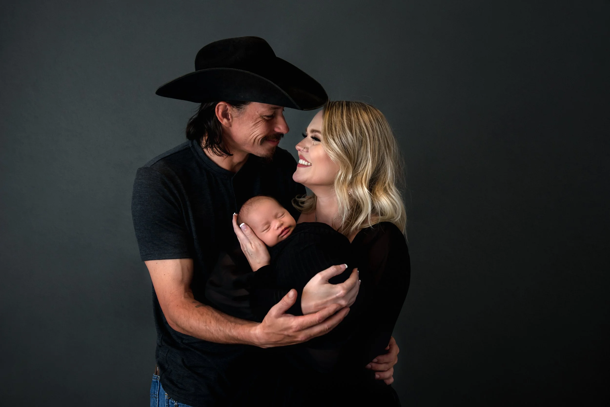 A smiling couple holding a newborn baby against a dark background. The man is wearing a black cowboy hat and black shirt, and the woman has blonde hair and a black top. They are looking at each other affectionately.