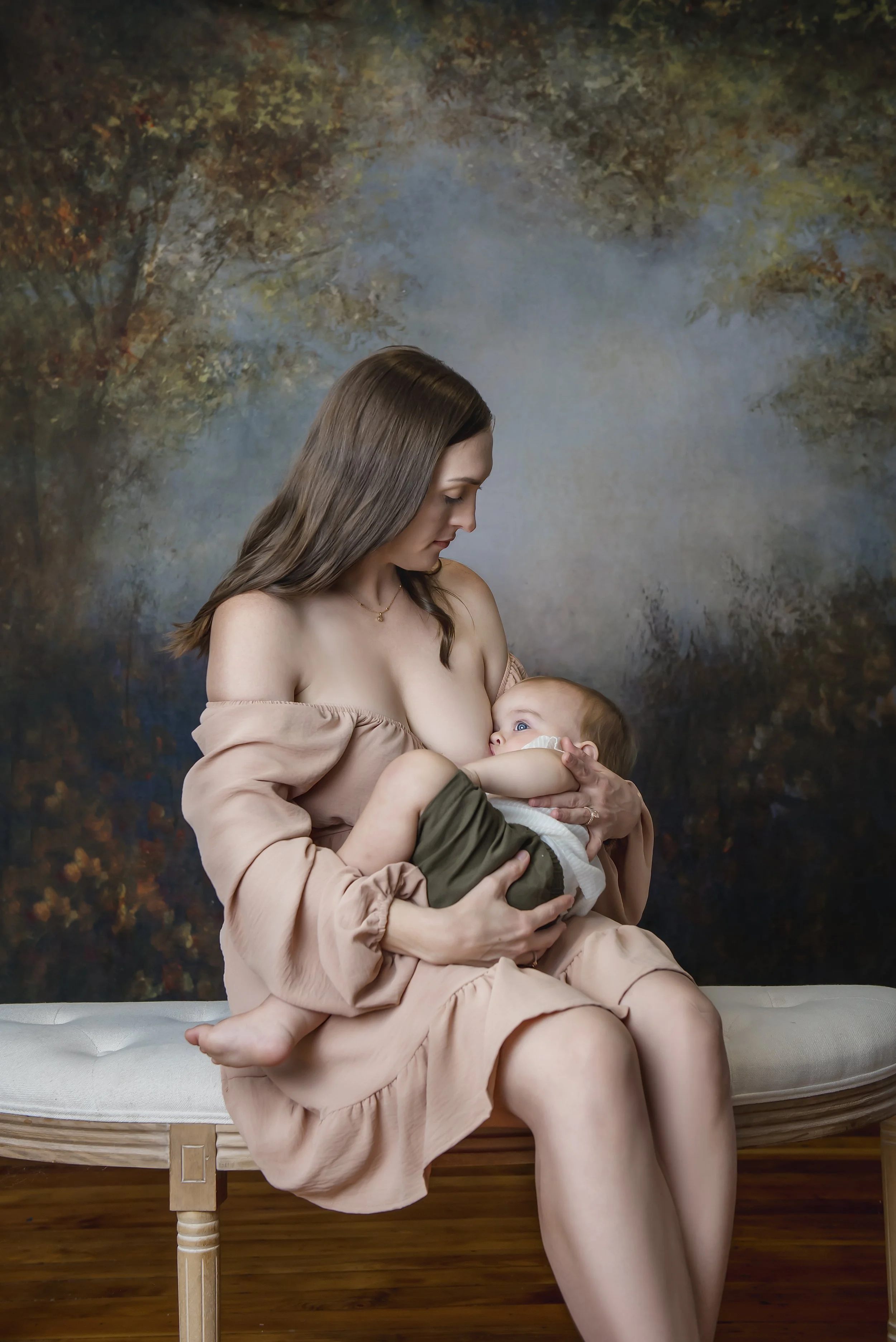 A woman breastfeeding a baby while sitting on a cream-colored bench with a wooden frame, against a painted backdrop of trees and sky.