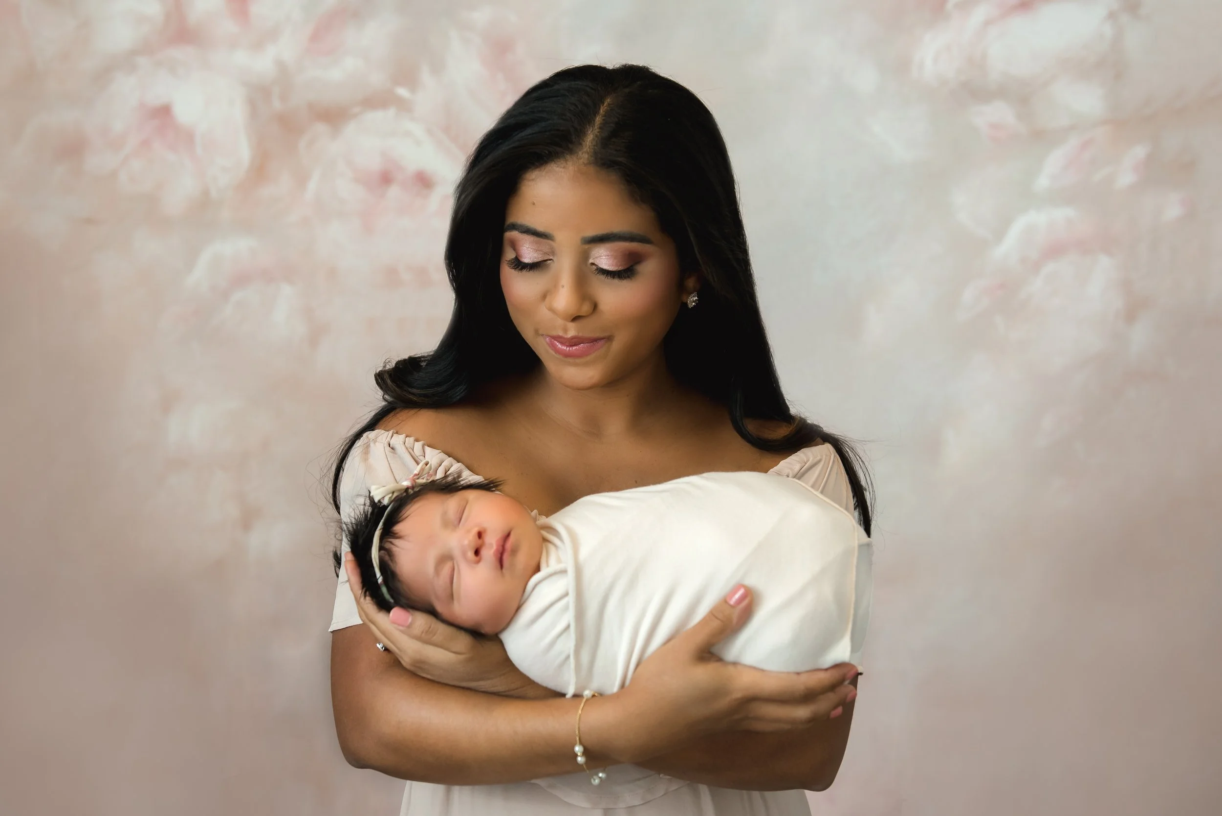 A woman holding a sleeping newborn baby wrapped in a cream blanket, with a gentle smile and closed eyes, against a soft pink background in our Spring Branch studio.