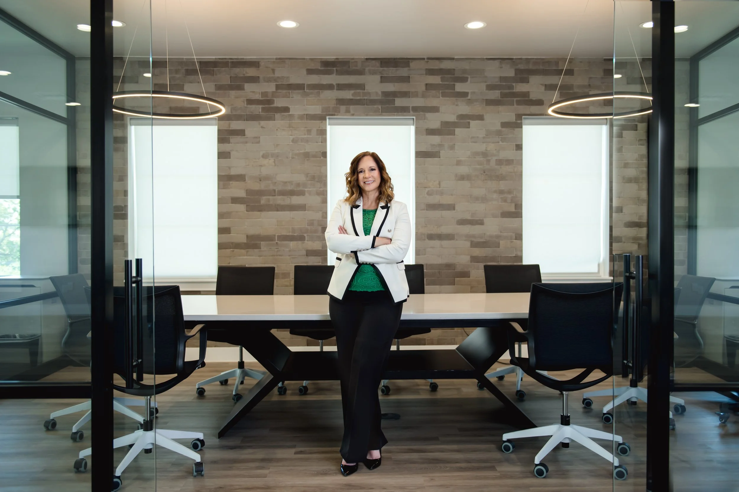 Woman standing in a conference room with a large table and black office chairs, with brick wall and frosted windows in the background.