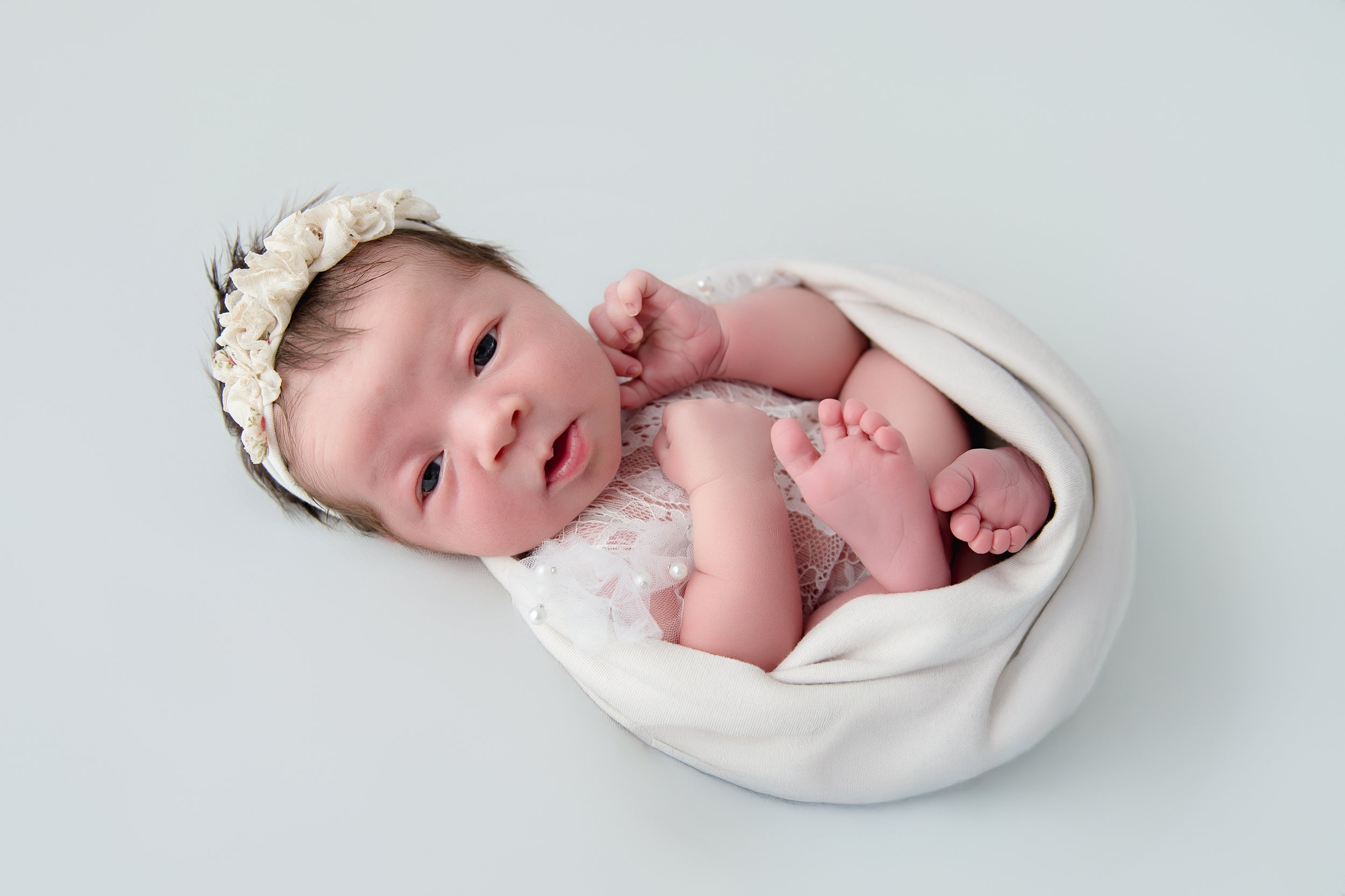 Adorable newborn baby girl with a floral headband, lying on a white blanket, in a delicate lace outfit.