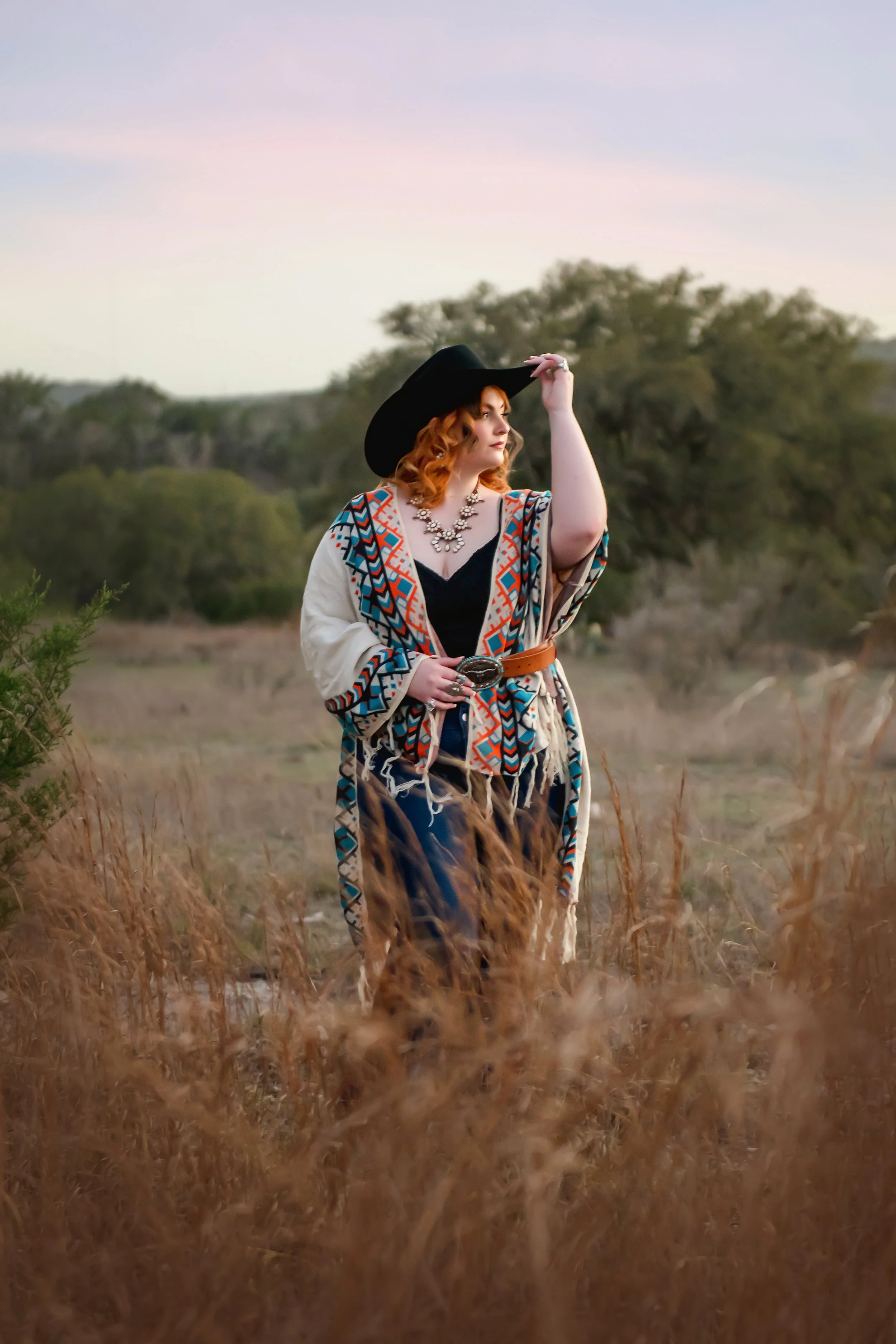 A woman with red hair, wearing a black hat, necklace, and colorful patterned shawl, standing in a field of tall grass during sunset.