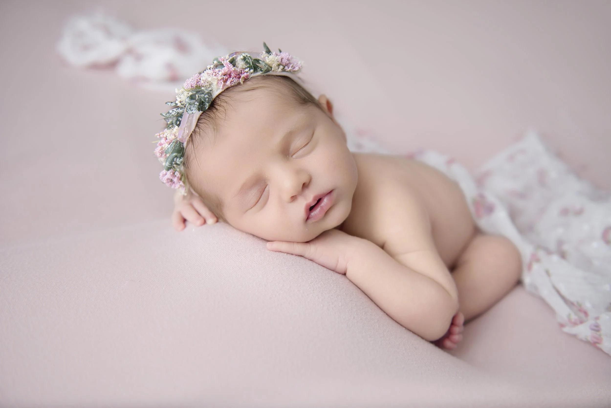 A sleeping baby lying on a soft pink surface, wearing a floral headband and a matching cloth.